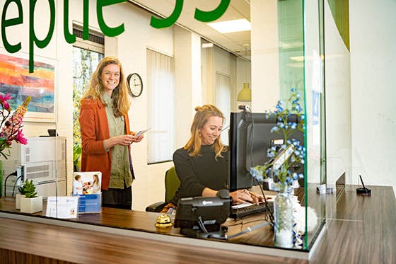 Twee vrouwen bij de receptie van de afdeling psychiatrie van het UMC Utrecht, één achter de computer en de ander staand met een document, beide glimlachend