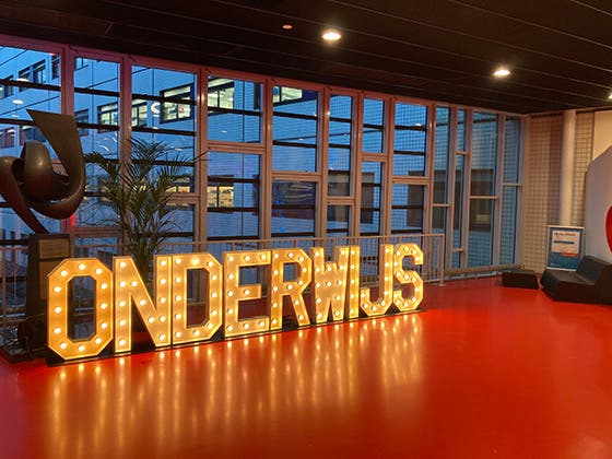 Grote verlichte letters die "ONDERWIJS" spellen in een modern interieur met rode vloer en grote ramen