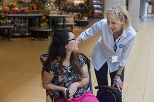 Een medewerker helpt een glimlachende patiënte in een rolstoel in de gang van het UMC Utrecht, met een bloemenwinkel op de achtergrond