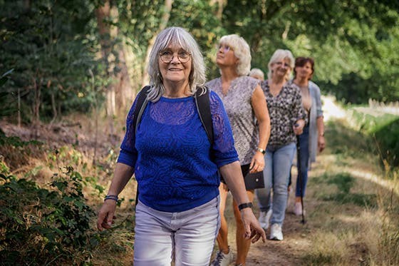 Oudere vrouw met grijze haren loopt voorop tijdens een wandeling met een groep vriendinnen in een bos.