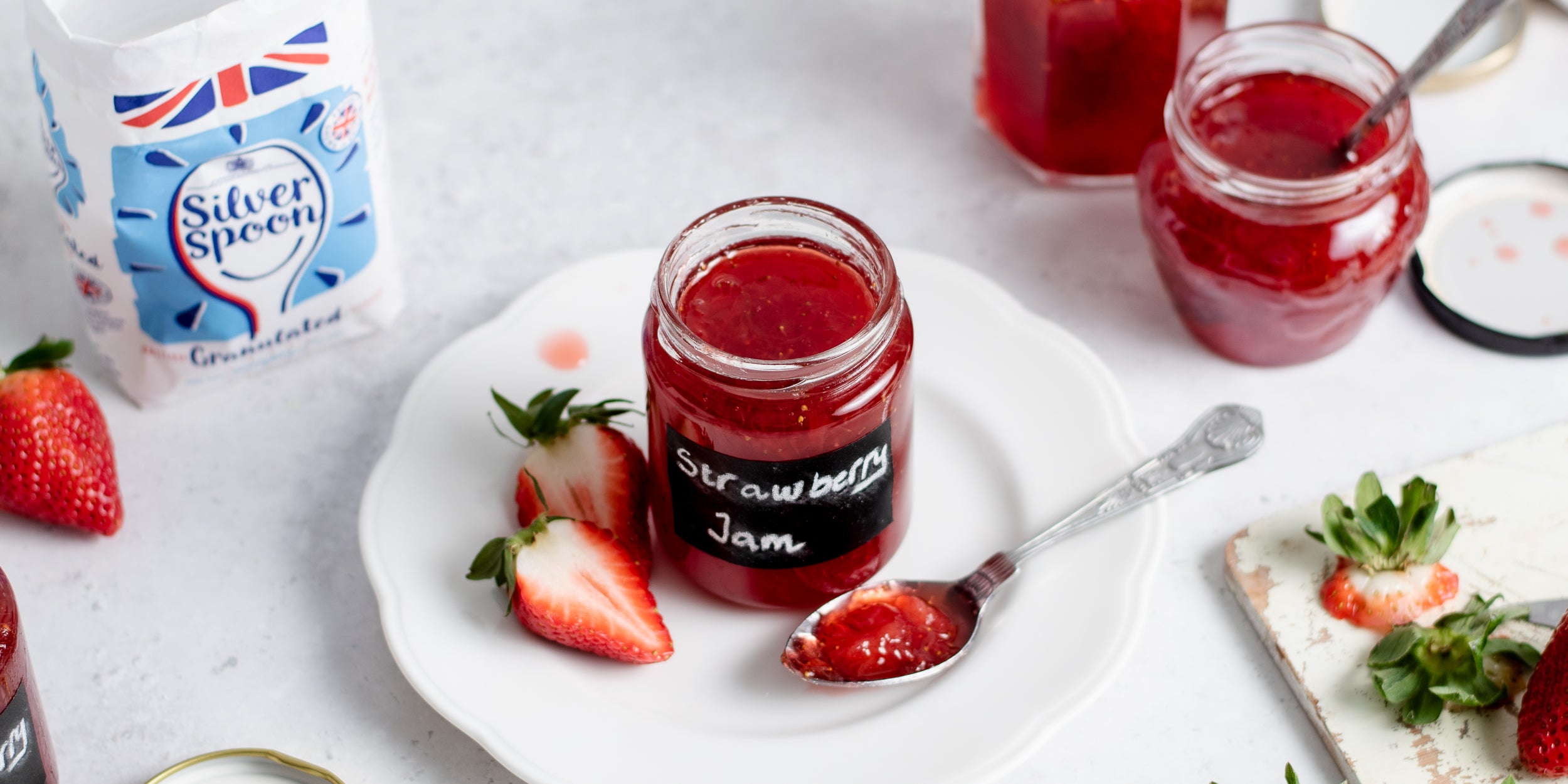 Strawberry Jam in a jam jar, with a spoon with a spoonful and sliced strawberries. Pack of Silver Spoon Granulated Sugar in the background