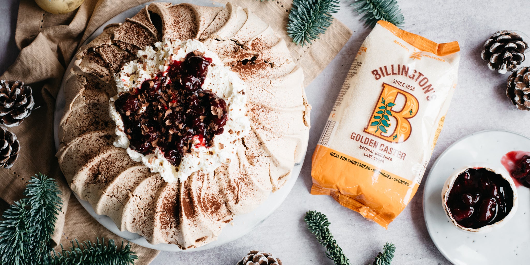 Black Forest Pavlova next to a bag of billingtons golden caster sugar, pine leaves and cones and a bowl of cherry kirsch