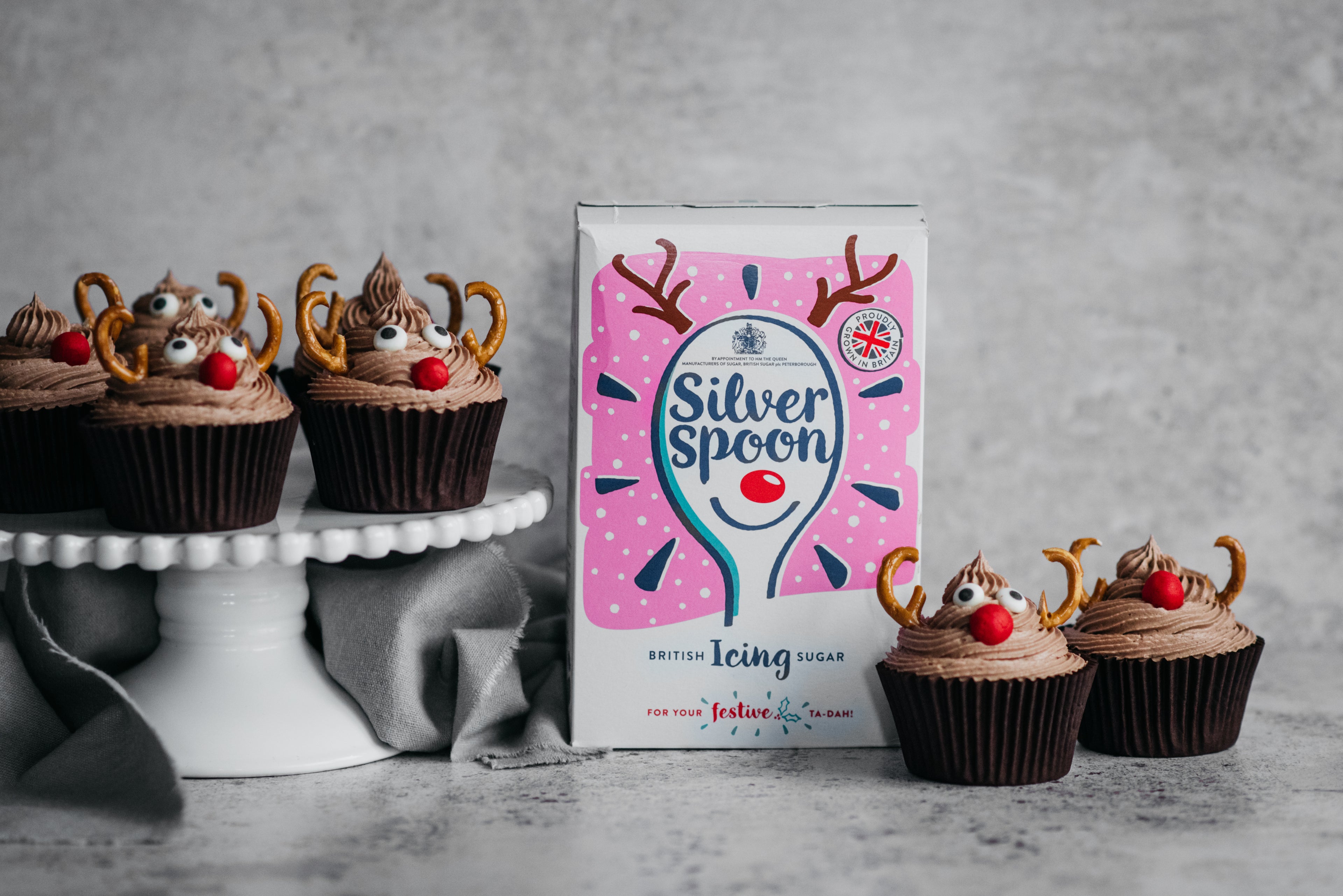 Reindeer Cupcakes on a cake stand, next to a box of Silver Spoon icing sugar, decorated with pretzel antlers and red nose sweets