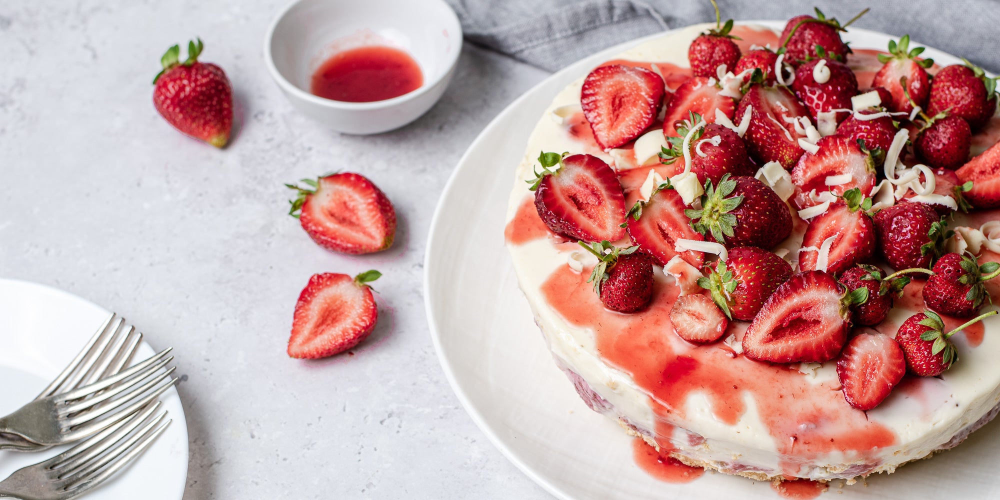 White Chocolate & Strawberry Cheesecake next to slices of strawberries and a small dish of strawberry sauce