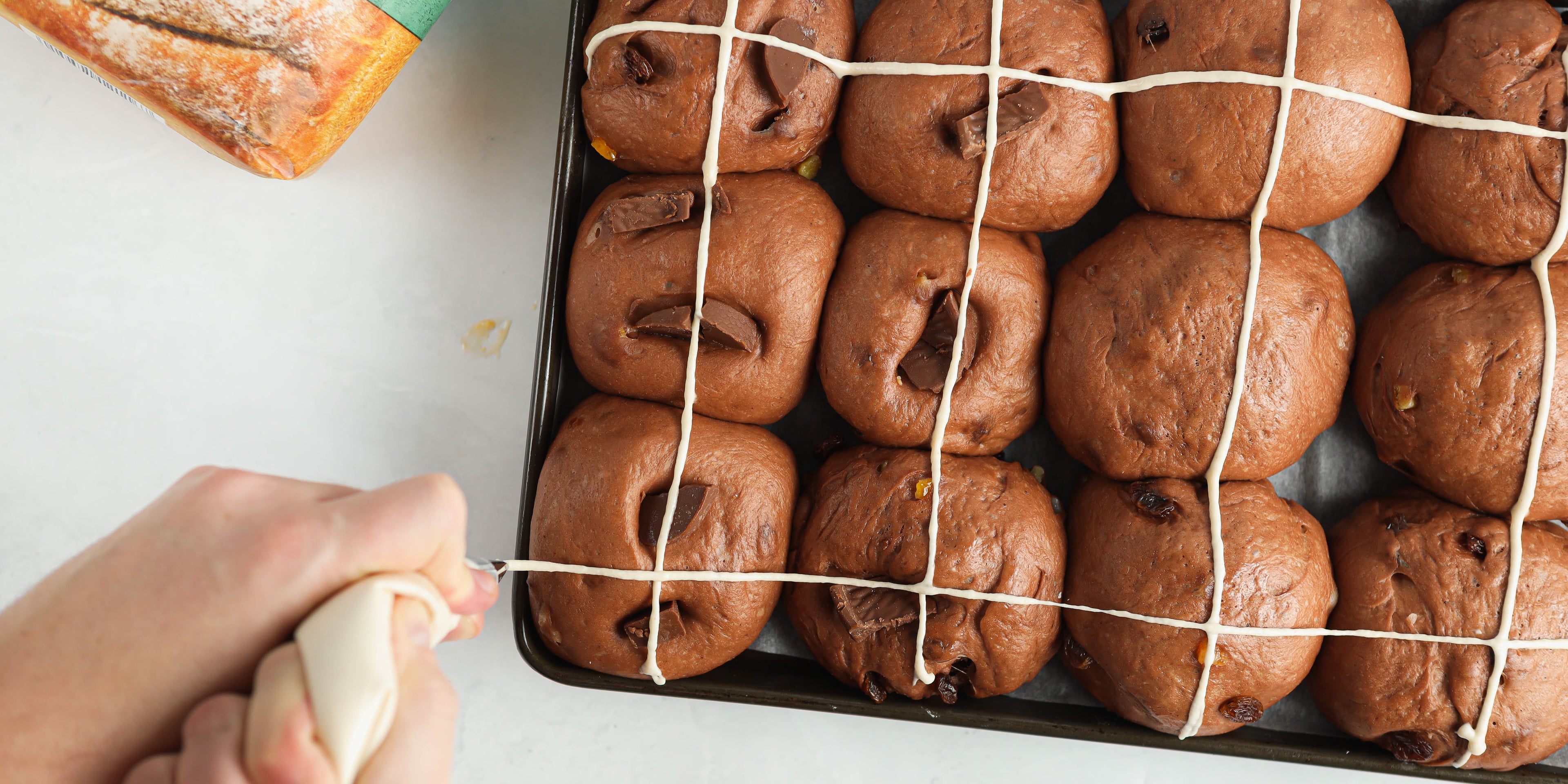 Top view of Chocolate Hot Cross Buns being piped with the traditional cross, with hands holding a piping bag