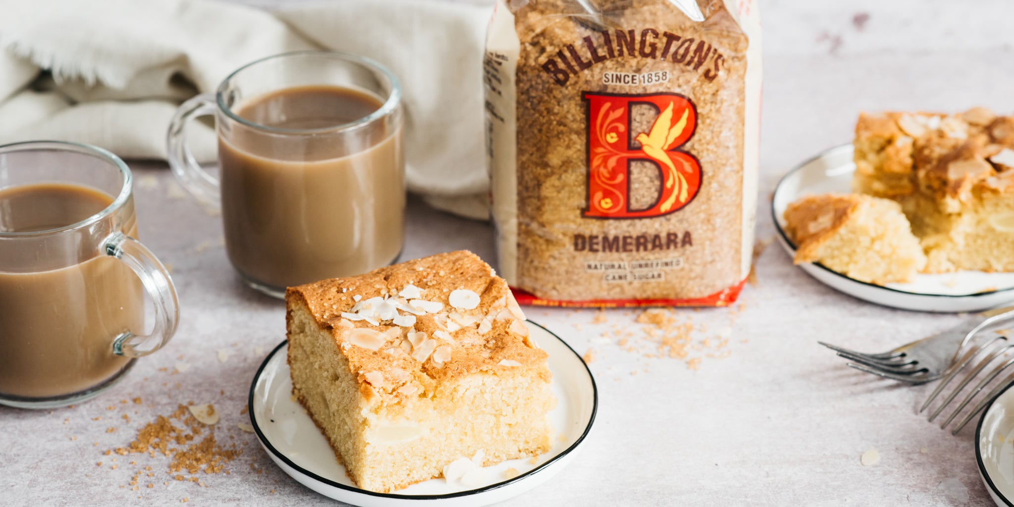 Square of cake on a plate beside 2 cups of coffee and a bag of sugar