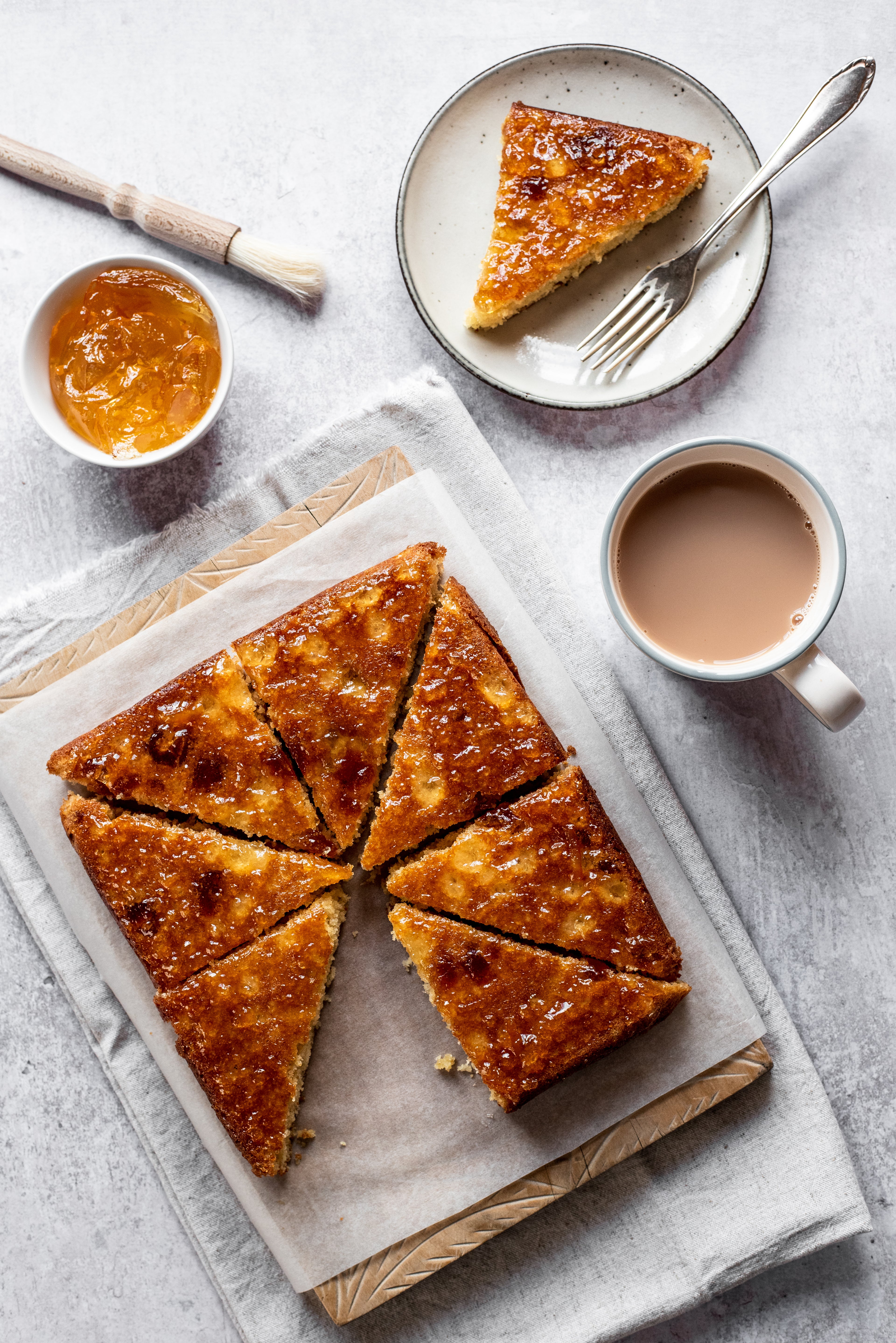 Top view of Marmalade Traybake sliced into pieces, on a sheet of baking paper, next to a cup of tea and a bowl of marmalade