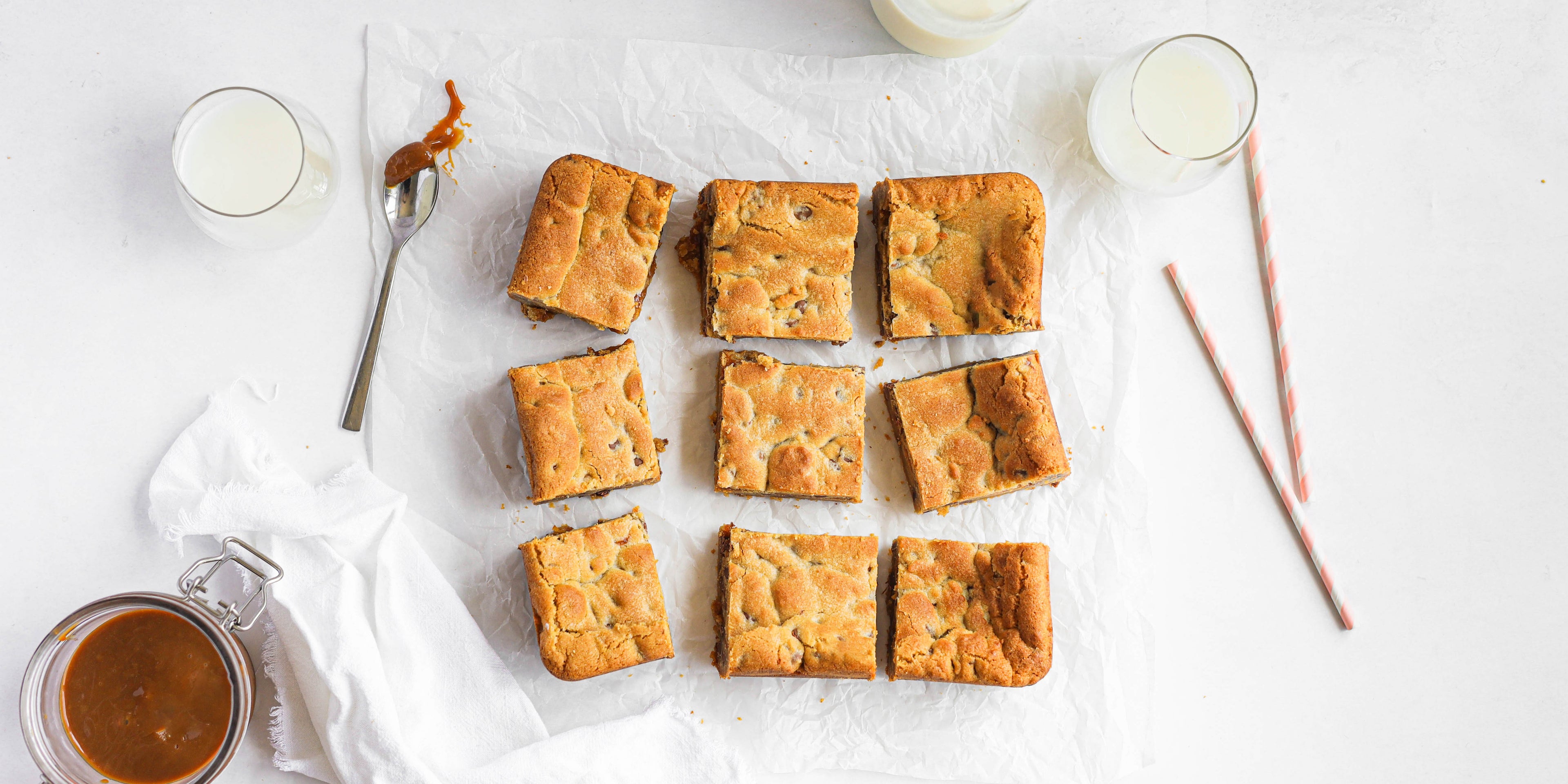 Salted Caramel Cookie Traybake cut into squares and lay on baking paper, next to a spoon with caramel sauce and glasses of milk