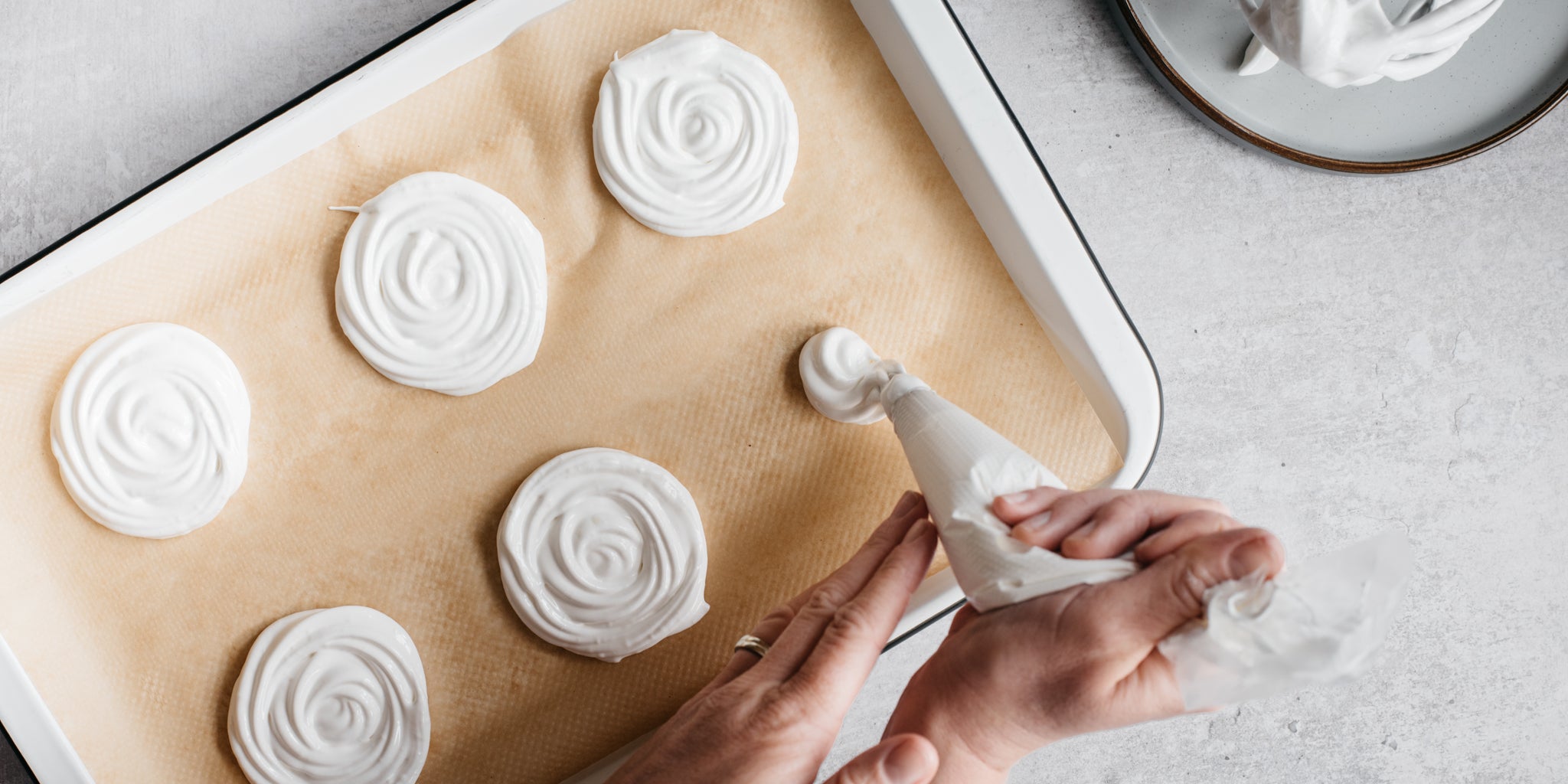 Two hands piping meringue into swirls on a tray lined with baking paper
