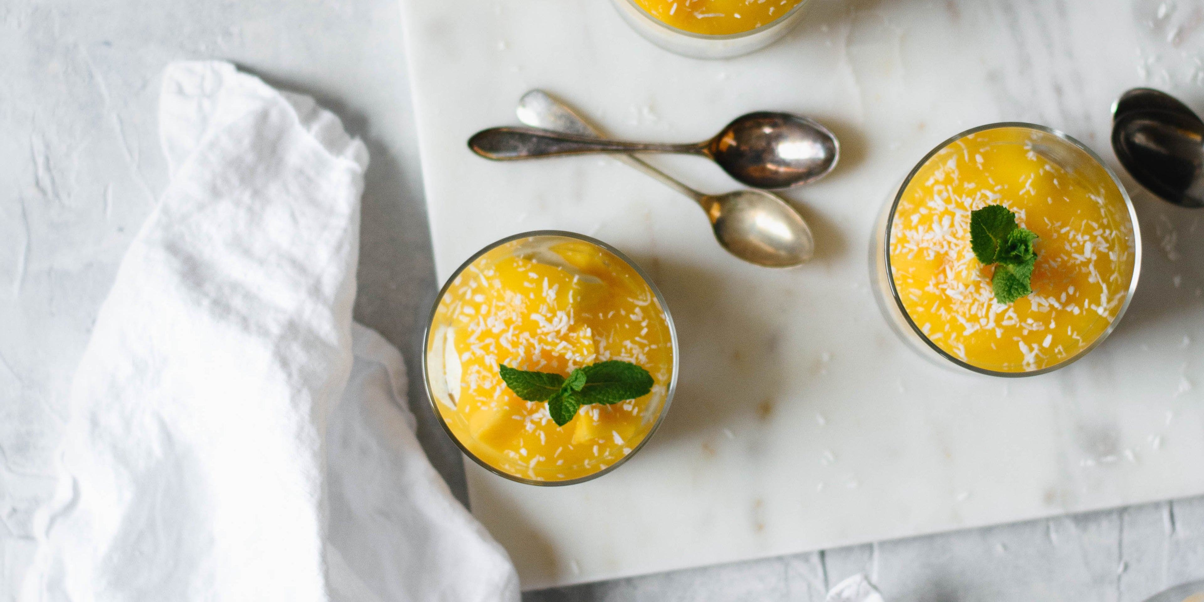 Overhead of small glasses of mango syllabub with sprig of mint on top and two spoons