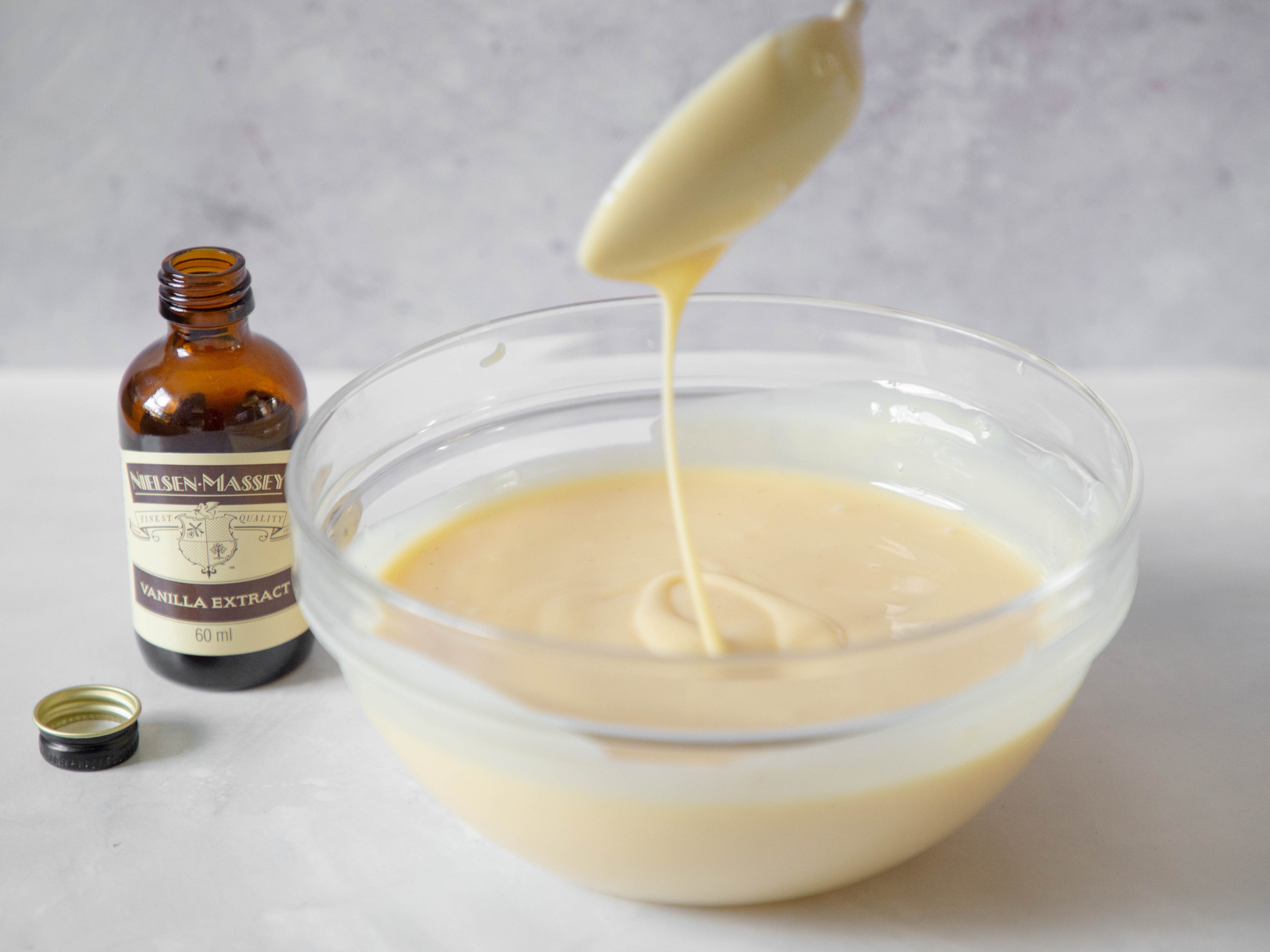 Glass bowl full of custard, with a spoon drizzling the custard above it. Bottle of vanilla extract to the left of the bowl with its lid off and infront of it