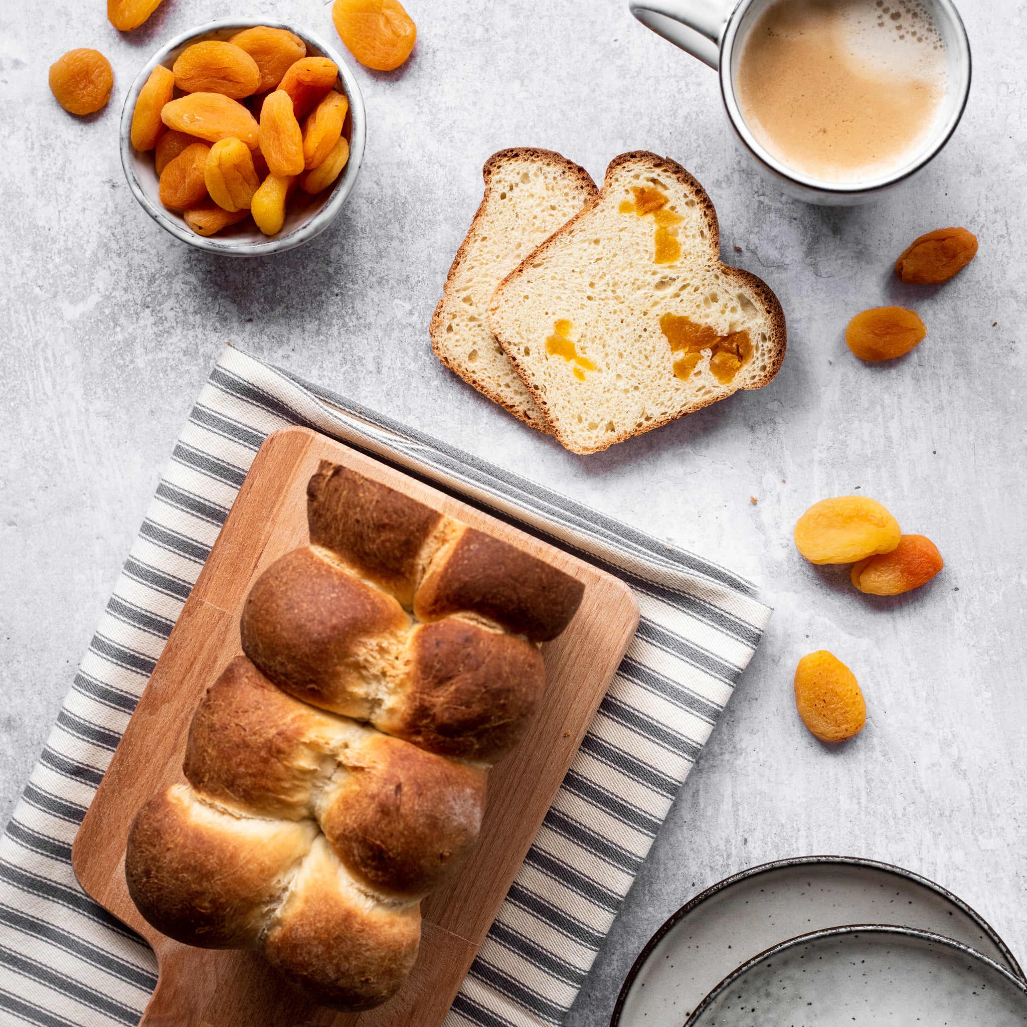 Overhead shot of brioche loaf with two slices cut in front, cup of tea and pot of apricots