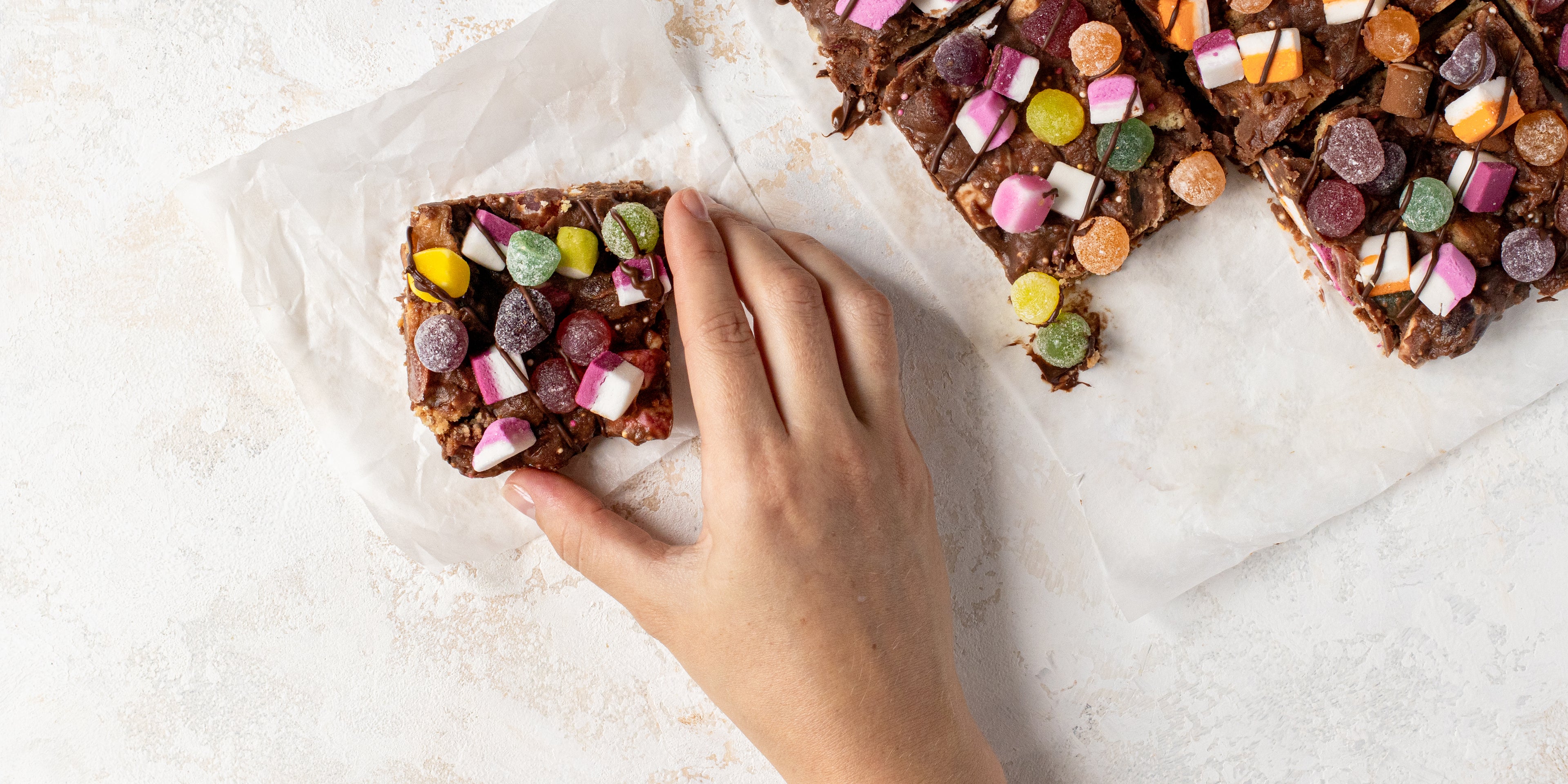 Top view of Sweetie Rocky Road lay on baking paper, with a slice cut out, and a hand reaching for a slice.