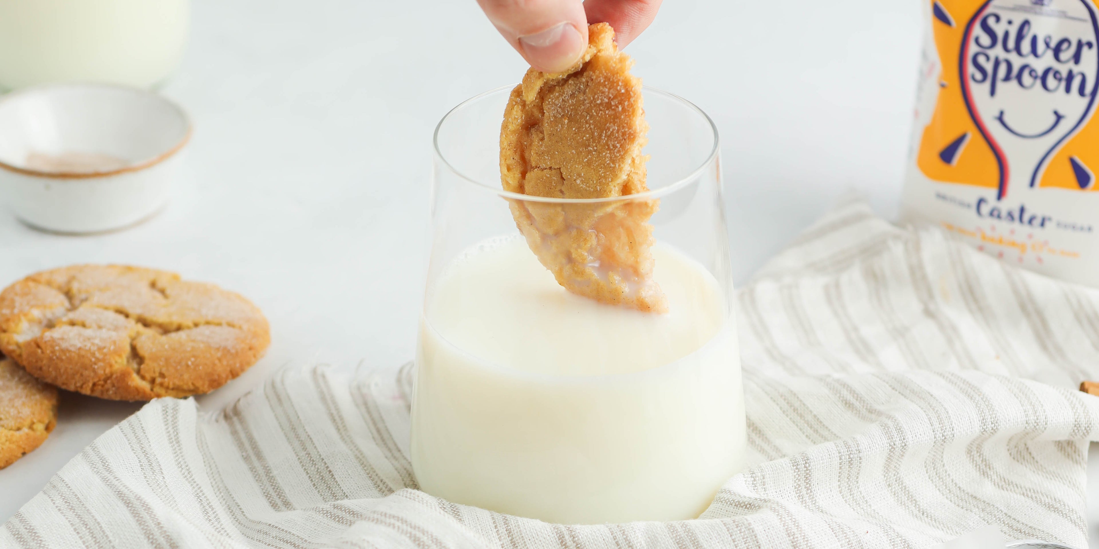 Close up of a Churro Cookies cut in half, being dunked into a glass of milk with a pack of Silver Spoon caster sugar in the background