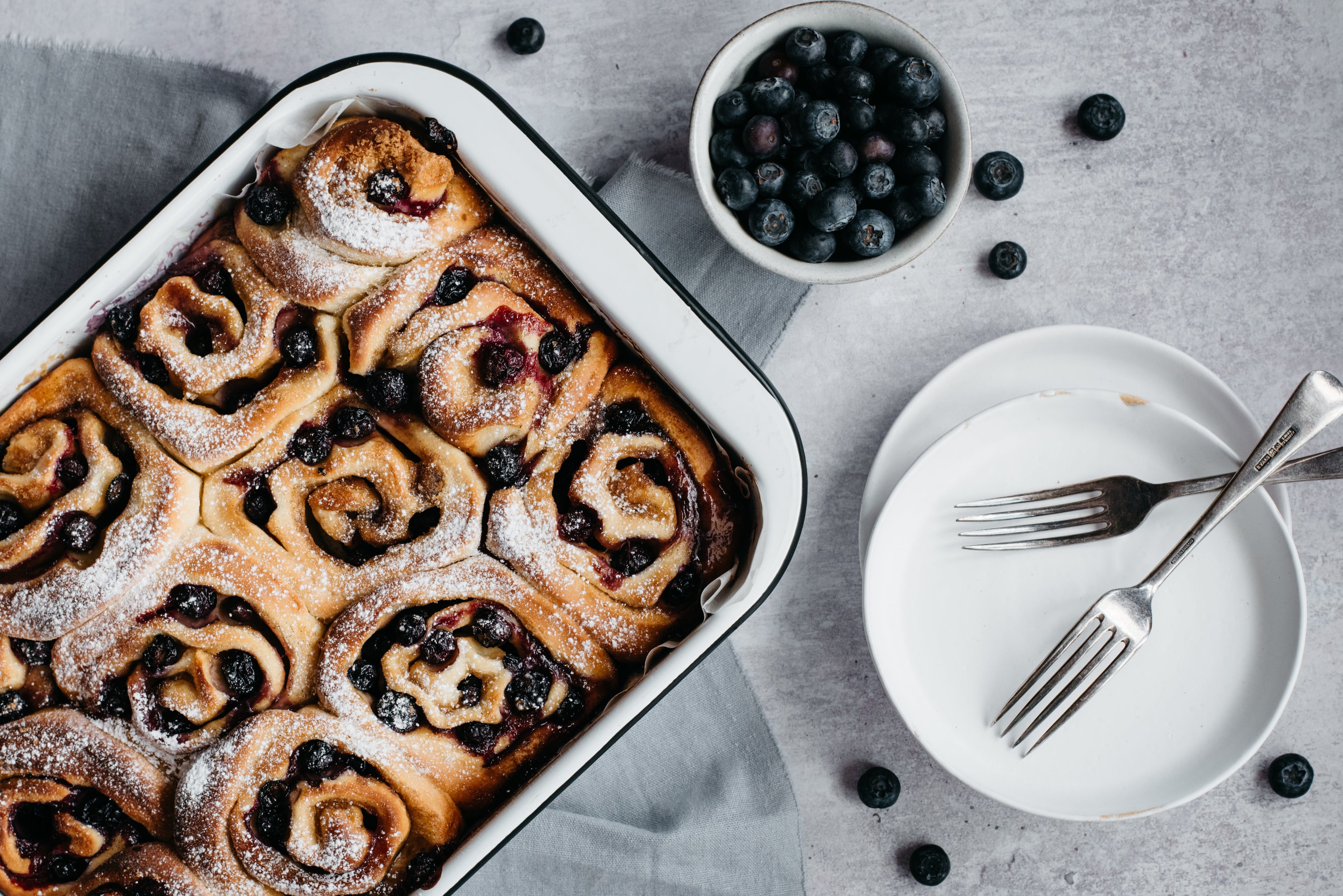 Tray of blueberry and vanilla rolls, topped with sugar, next to plates, cutlery and a bowl of blueberries