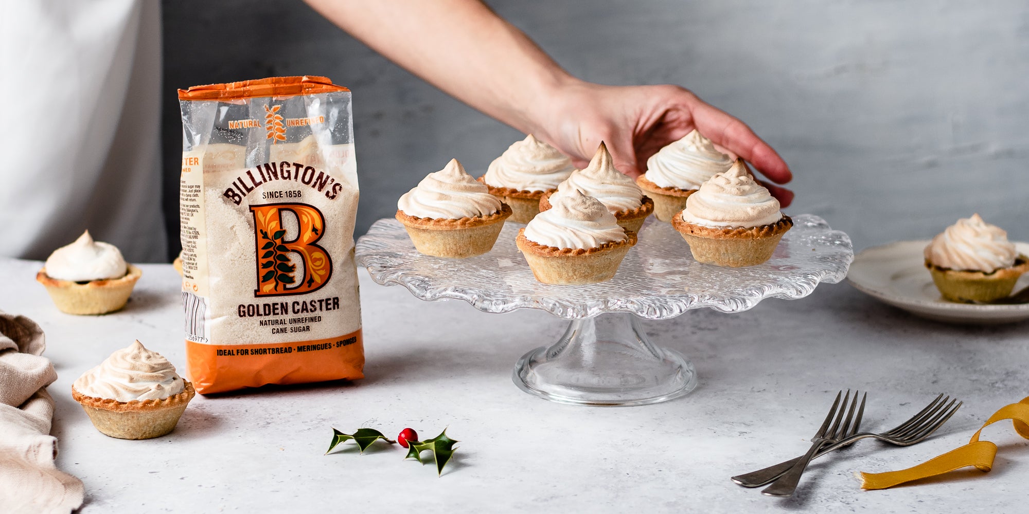 Meringue Topped Mince Pies on a glass cake stand with a hand reaching for one next to a bag of Billington's golden caster sugar