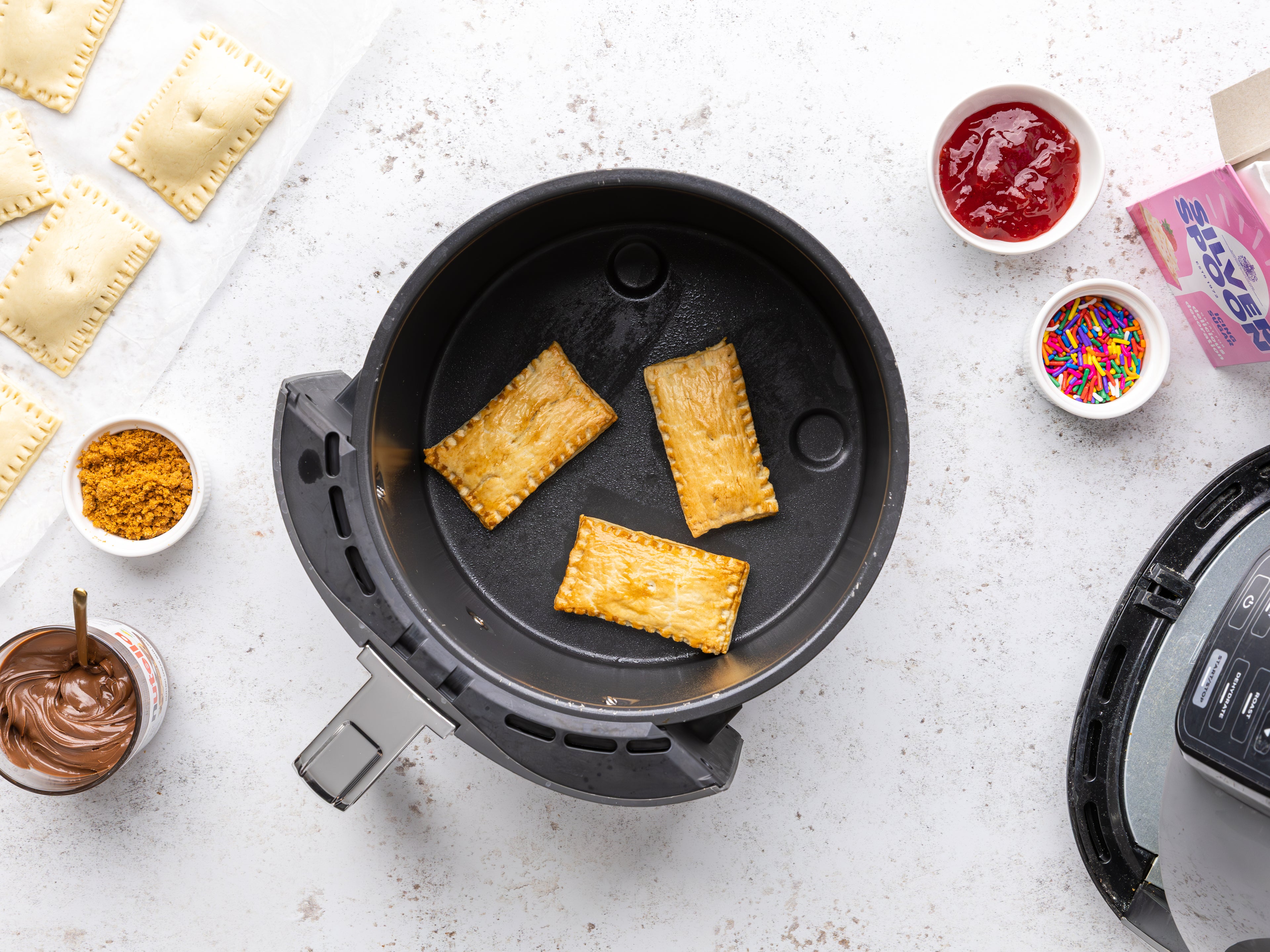 Pop Tarts in an air fryer surrounded by jam, sprinkles and open pack of icing sugar