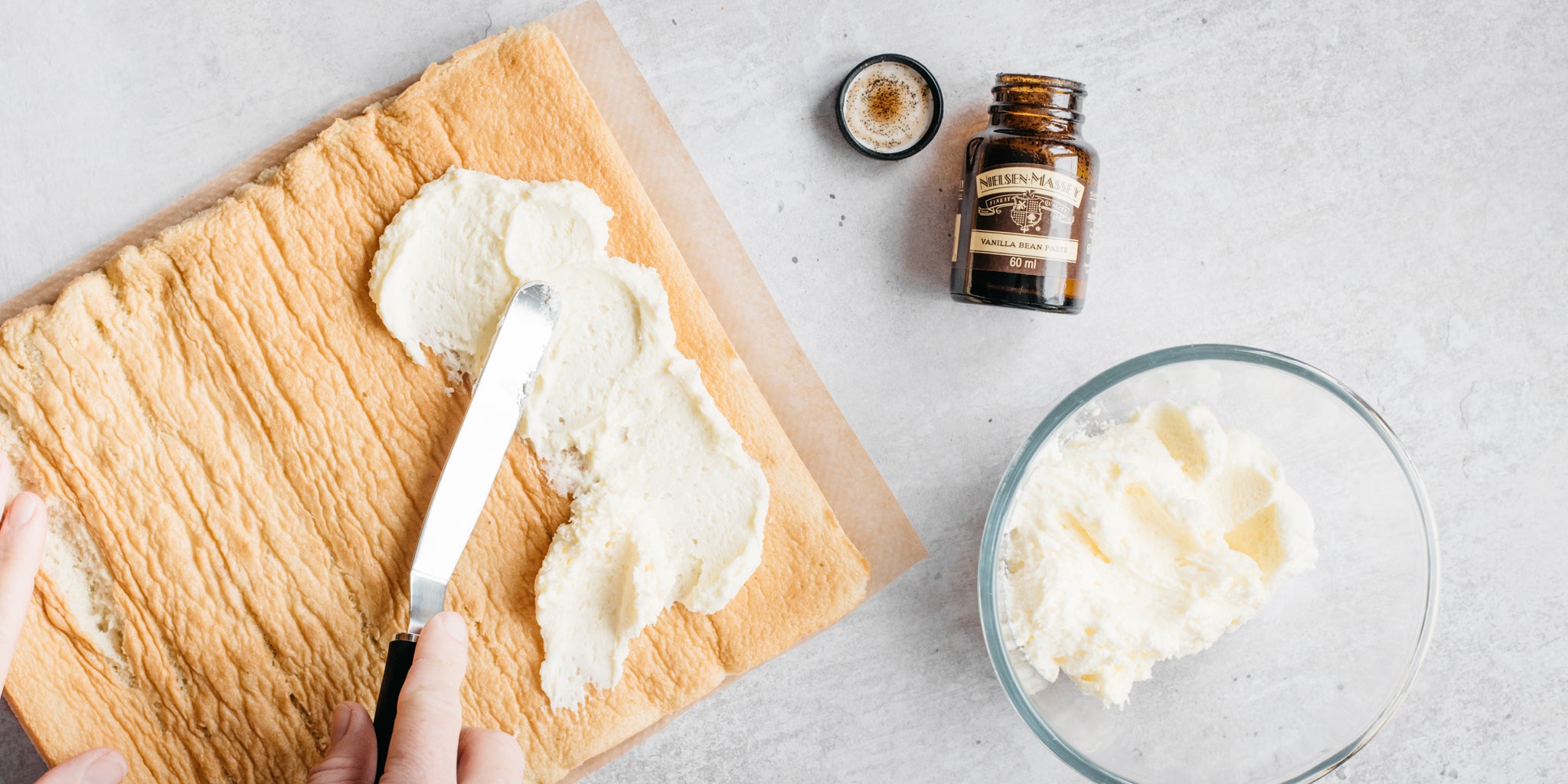 Top down view of a hand adding cream to a swiss roll next to a bottle of nielsen massey vanilla bean paste