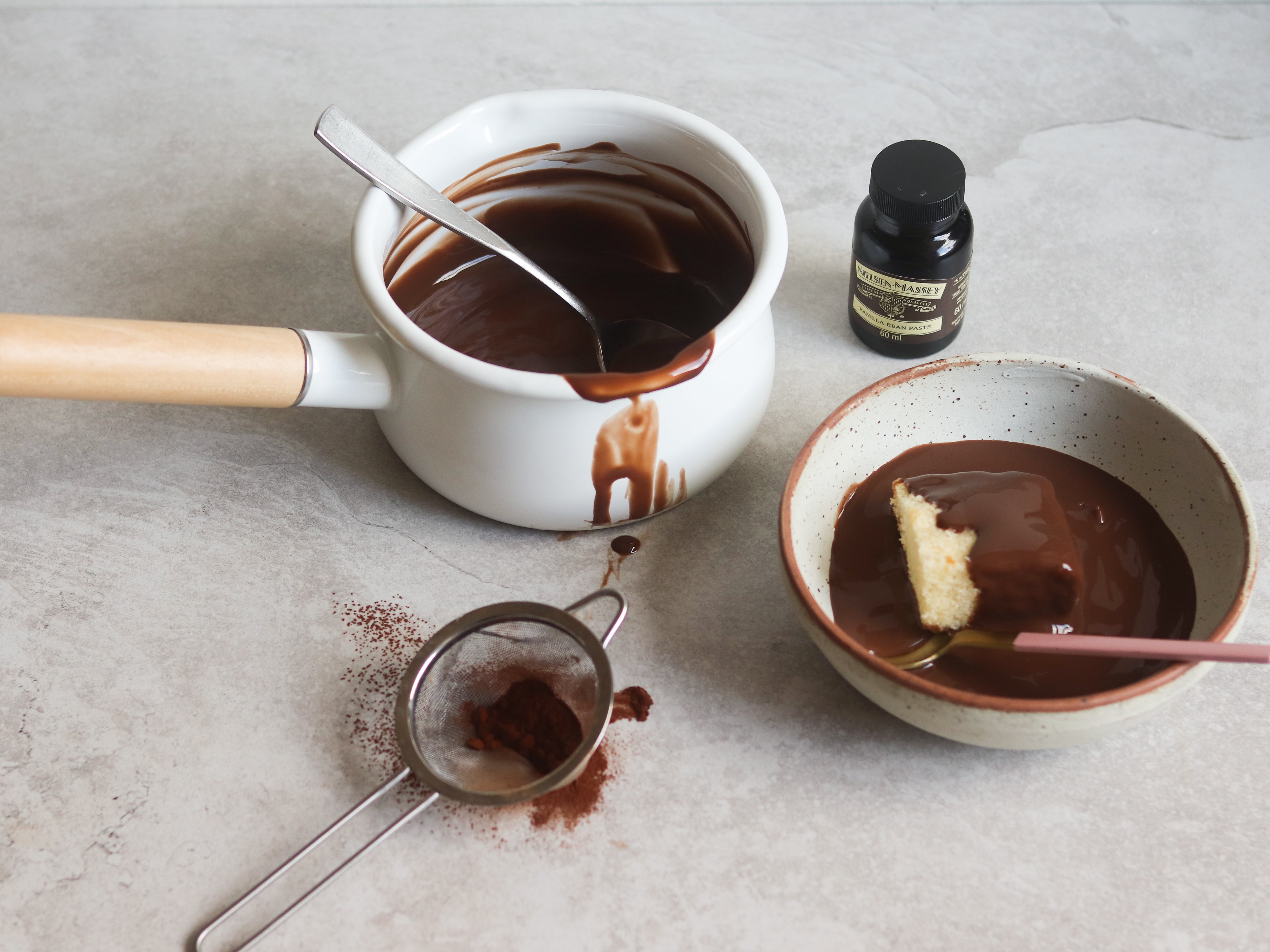 Saucepan of Chocolate Custard with drips down the side, next to a sieve with cocoa powder, and a bowl of sponge cake covered in Chocolate Custard with a spoon. Bottle of Neilsen-Massey Vanilla Extract in the background