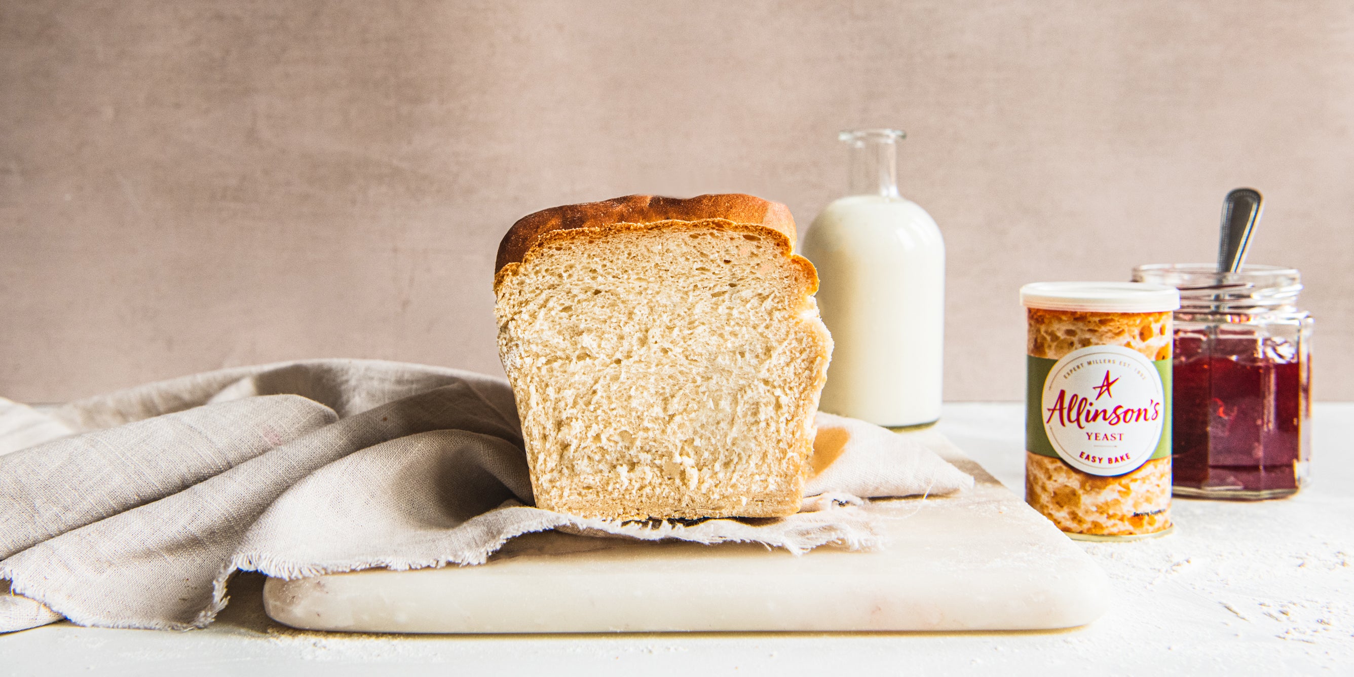 Shokupan Milk Bread with a slice cut off, showing the fluffy dough inside the loaf. Next to a jar of jam and a tin of Allinson's yeast