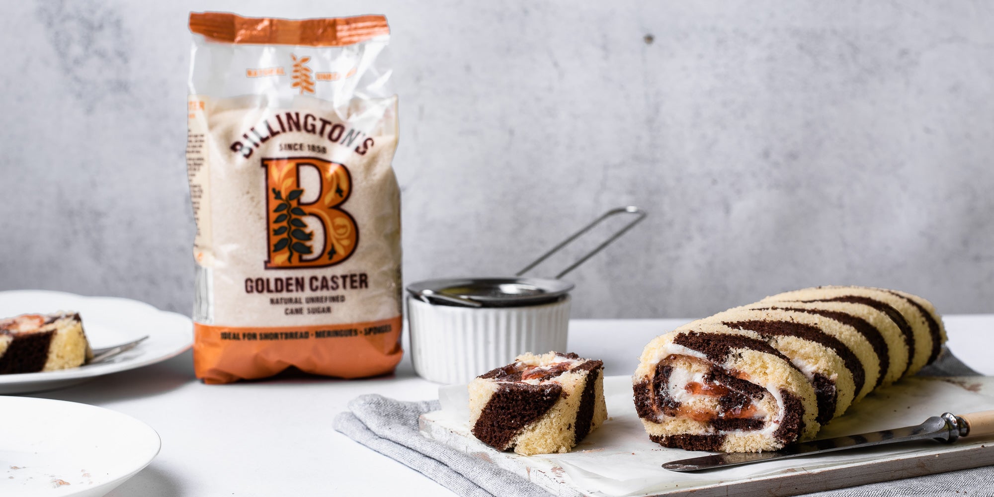 A sliced Chocolate & Vanilla Swiss Roll, next to a bag of Billington's Golden Caster sugar