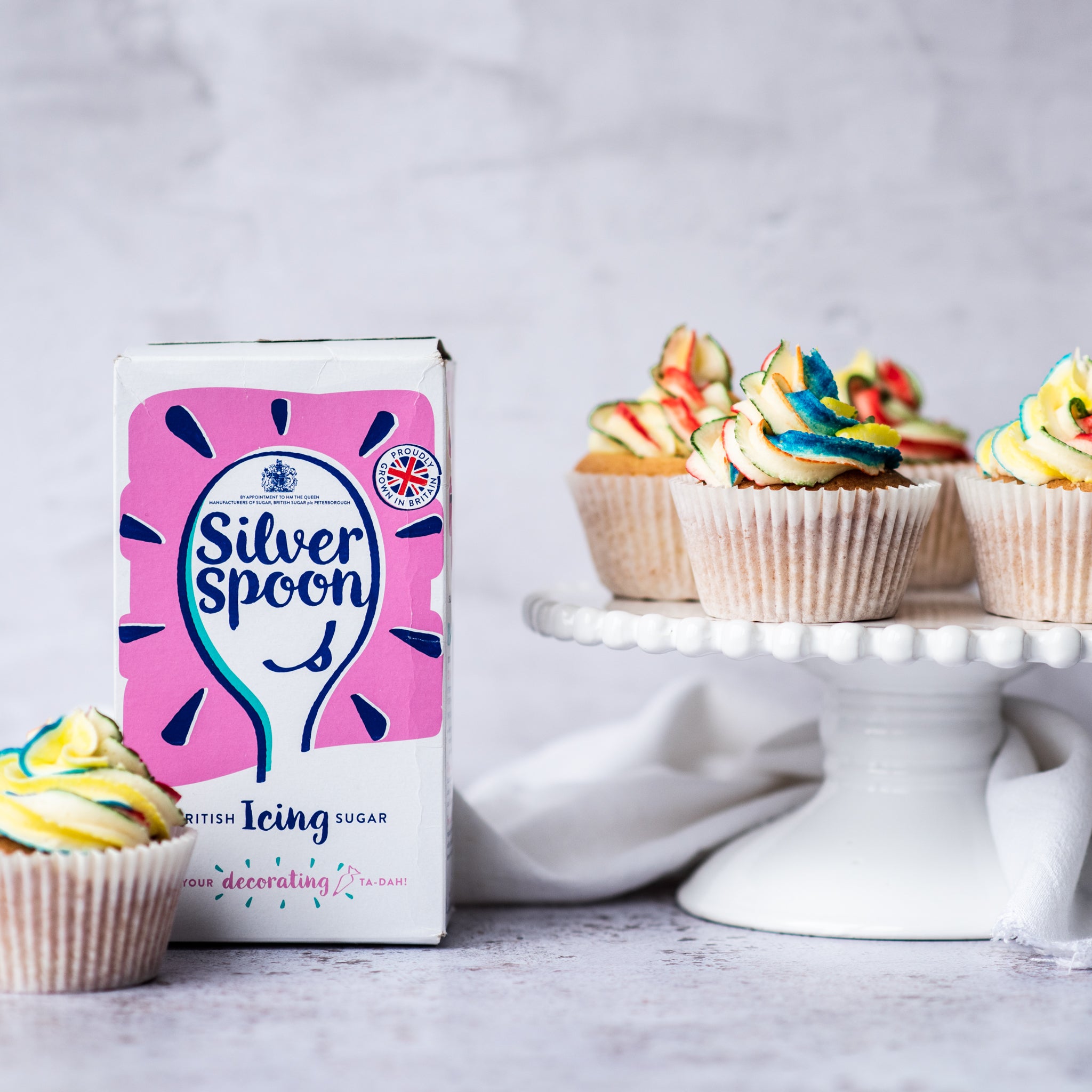 Cupcakes on stand with rainbow icing and box of icing sugar