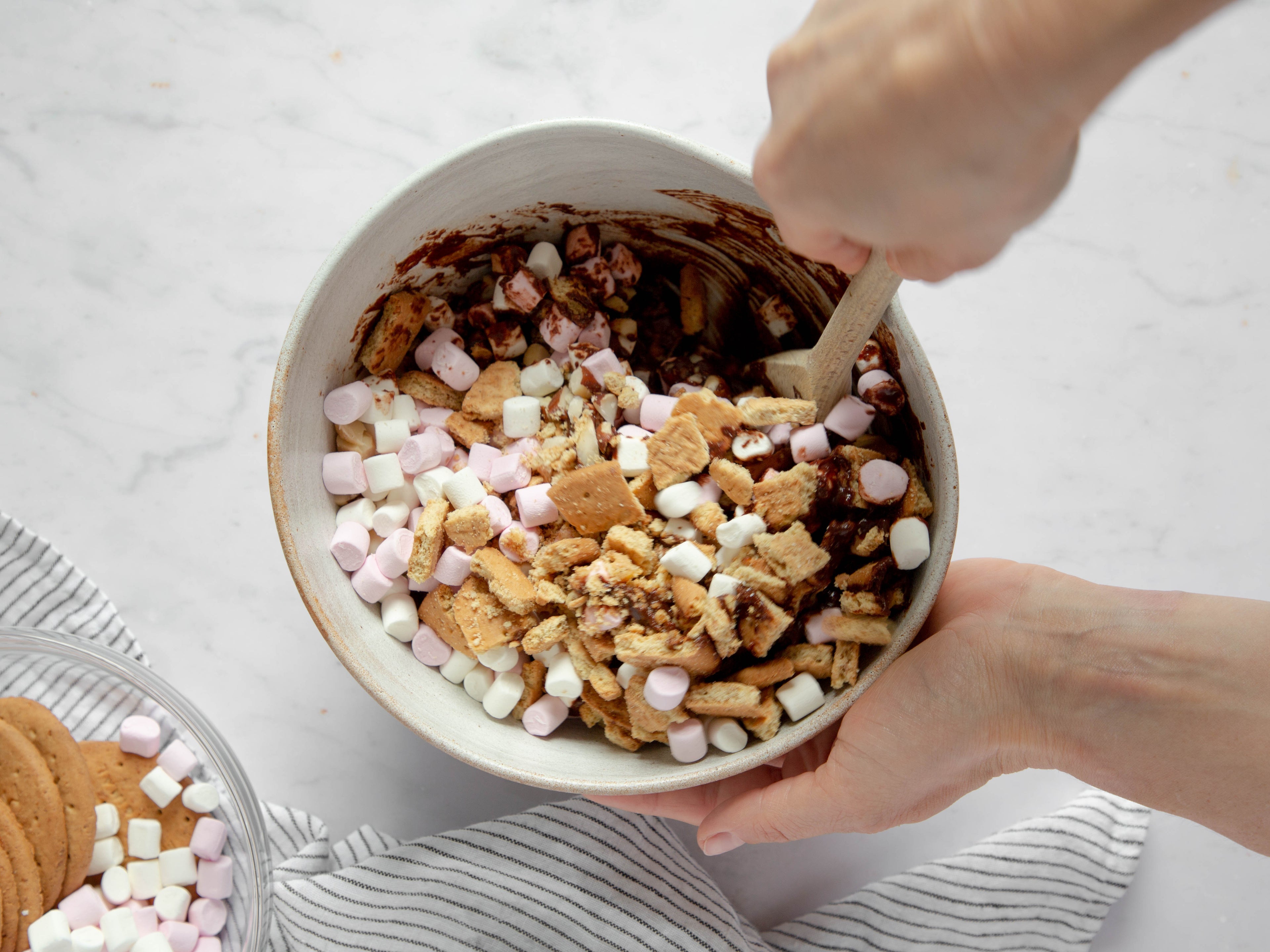 Hand holding a bowl and stirring rocky road ingredients in it with a wooden spoon. Striped tea towel in forefront with another bowl of ingredients