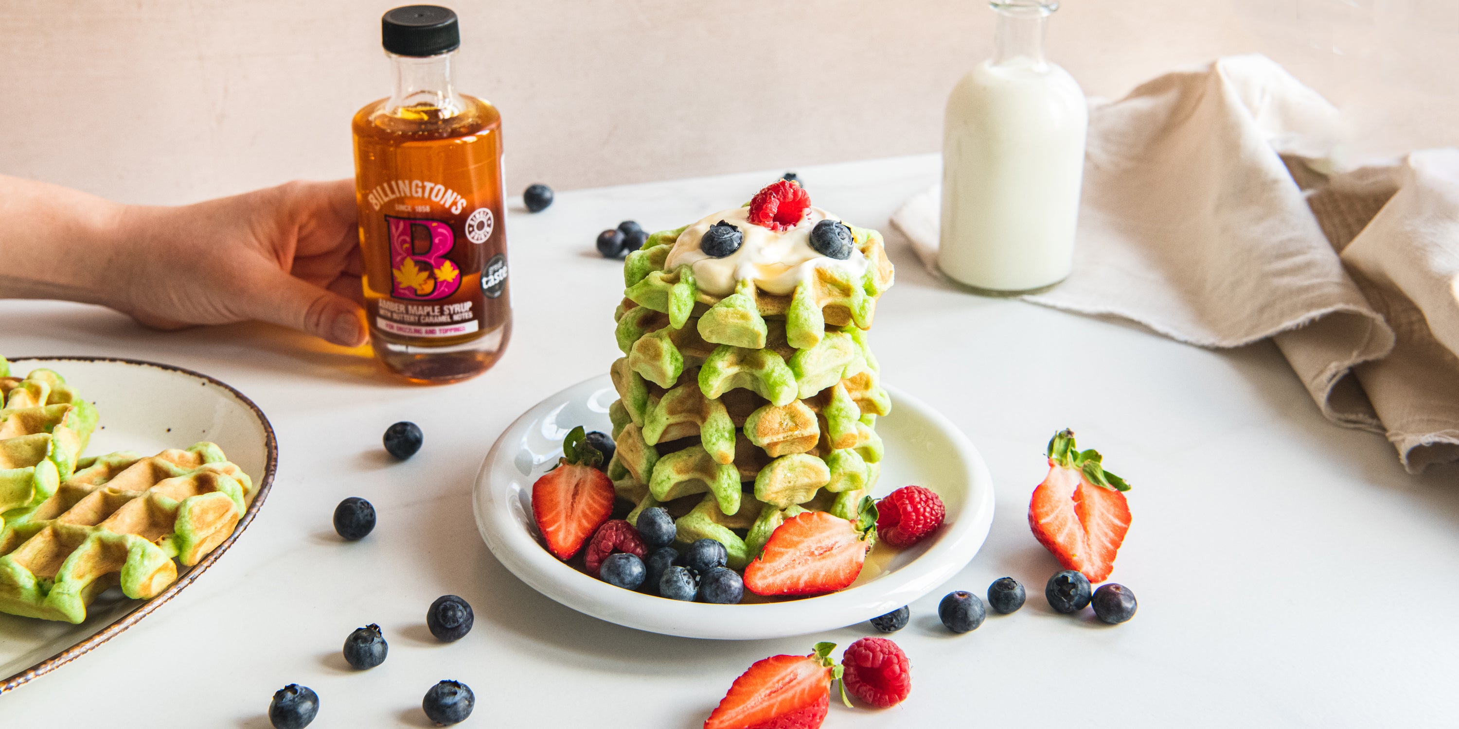 Pandan Waffles stacked with fresh berries, next to a hand holding a bottle of Billington's maple syrup and scattered blueberries