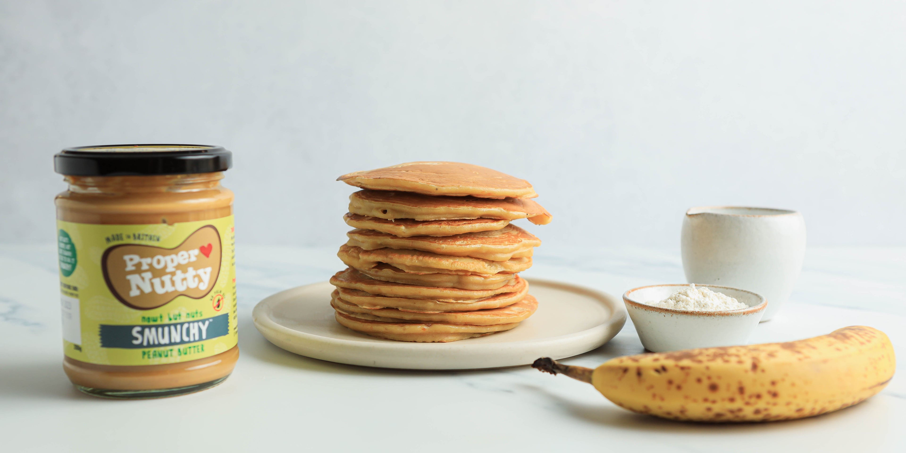 Stack of pancakes on a white plate next to a jar of peanut butter, banana and a small white bowl and cup