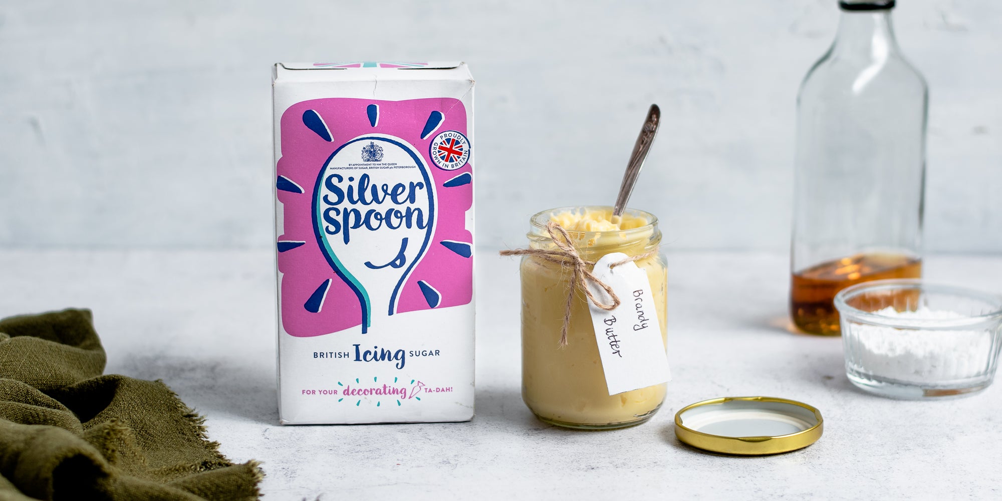 Glass jar of Brandy Butter with a handwritten notes, next to a glass bottle of brandy and a box of silver spoon icing sugar