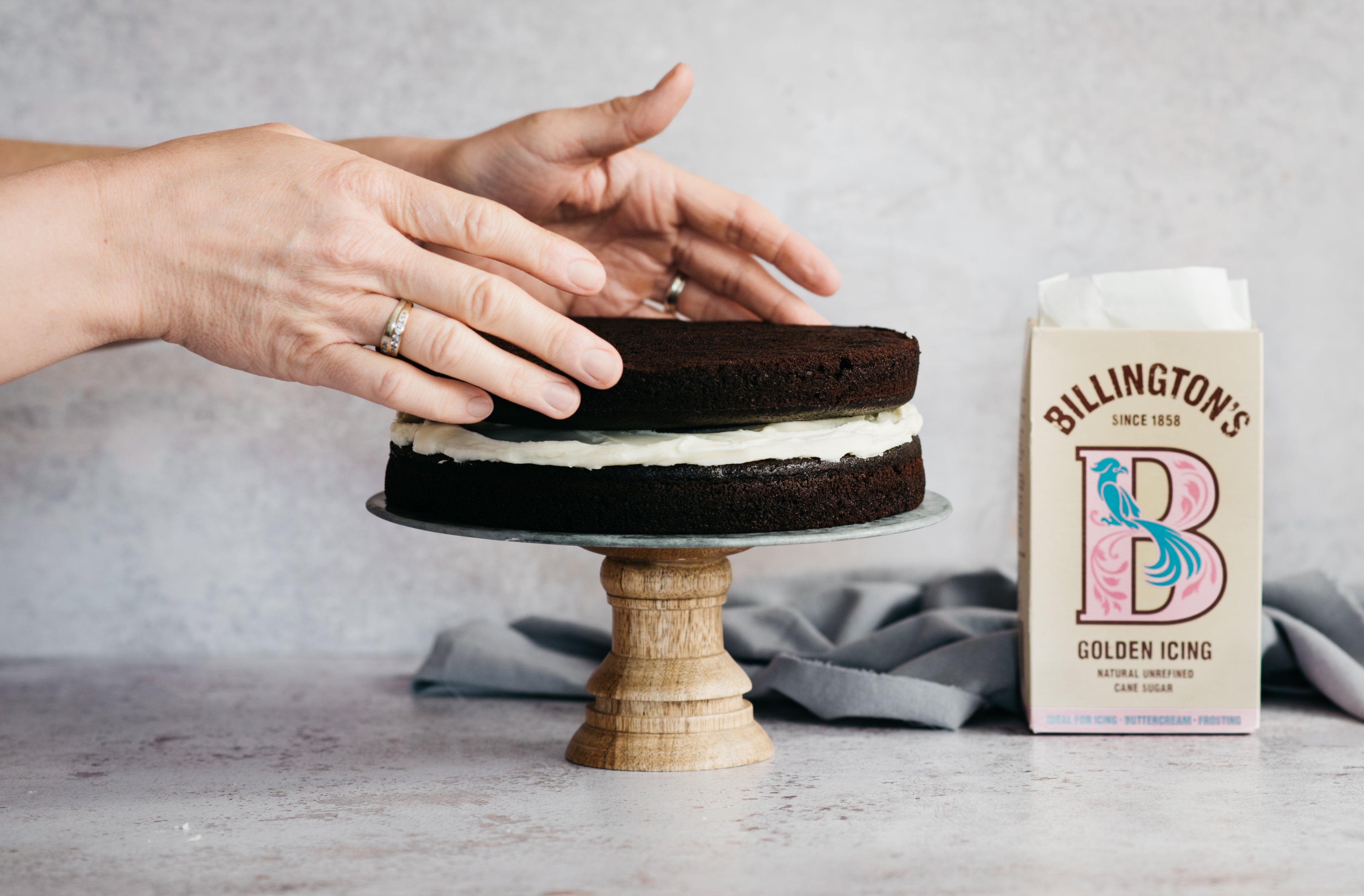 Hands layering Billington's Best Chocolate Cake between a layer of buttercream using Billington's Unrefined Golden Icing Sugar