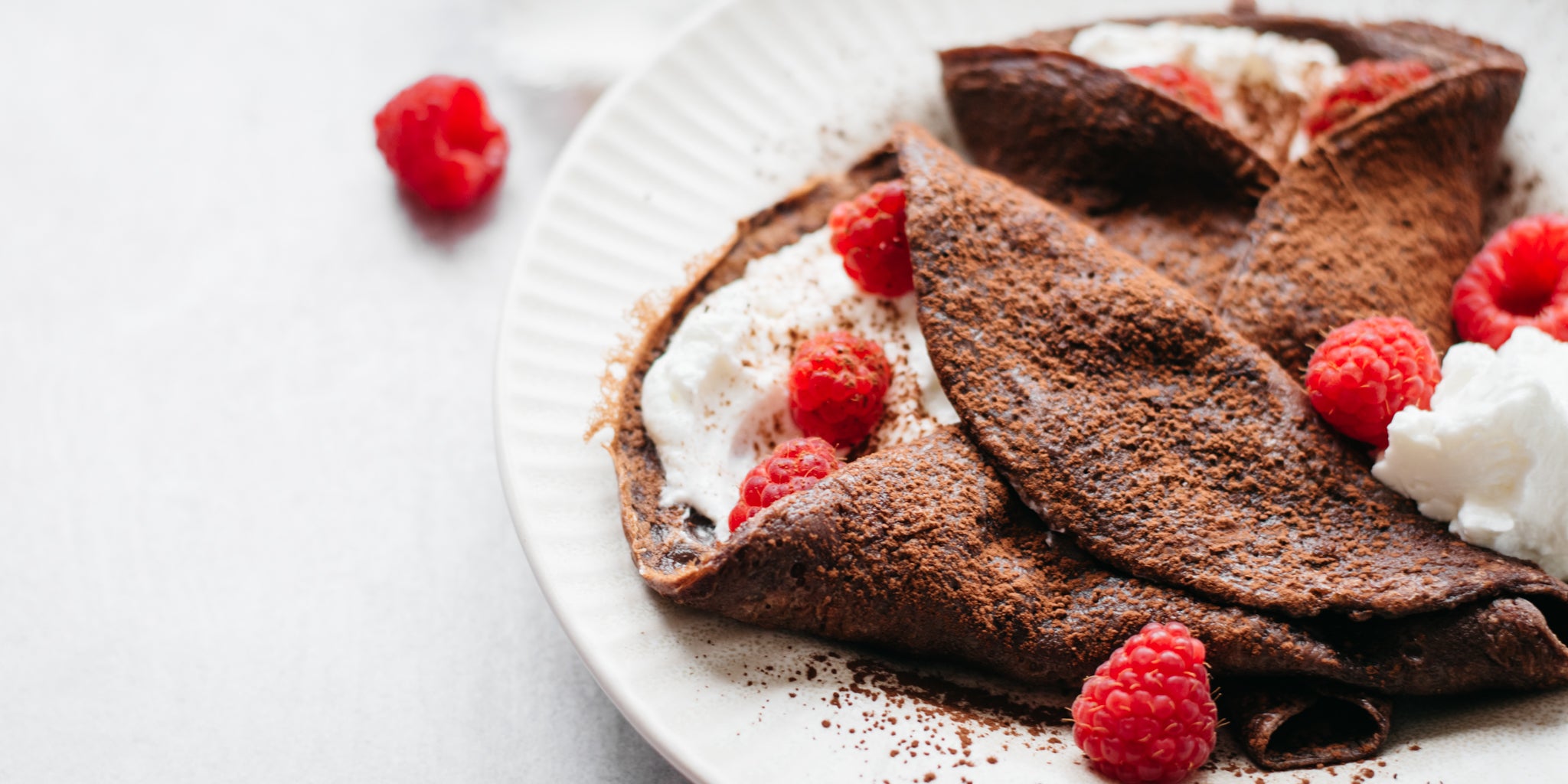 Close up shot of two chocolate crepes on a white plate surrounded by raspberries