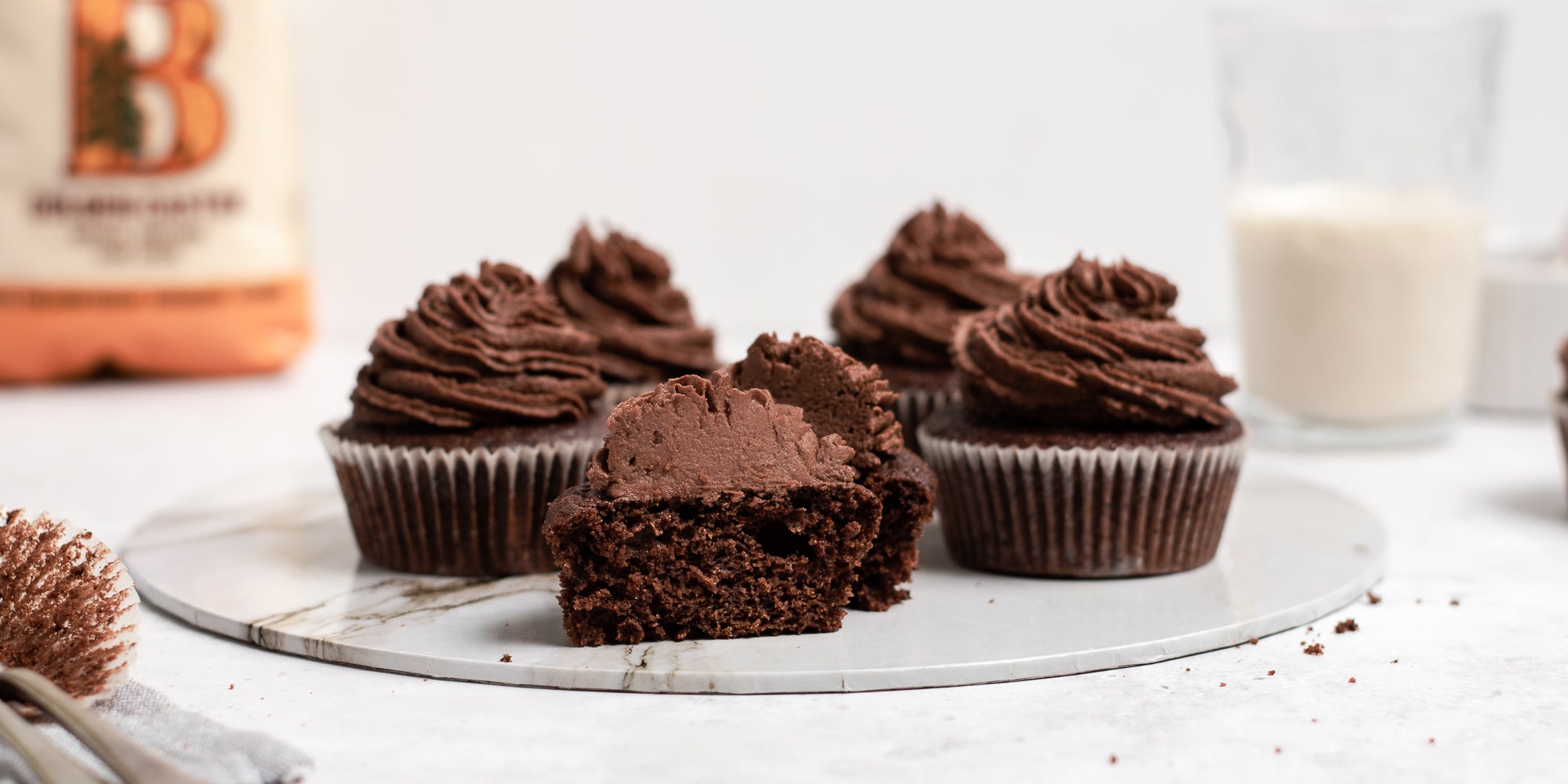Close up of Vegan Chocolate Cupcakes on a marble board, with a cupcake cut in half showing the centre. A pack of Billington's Golden Caster sugar in the background