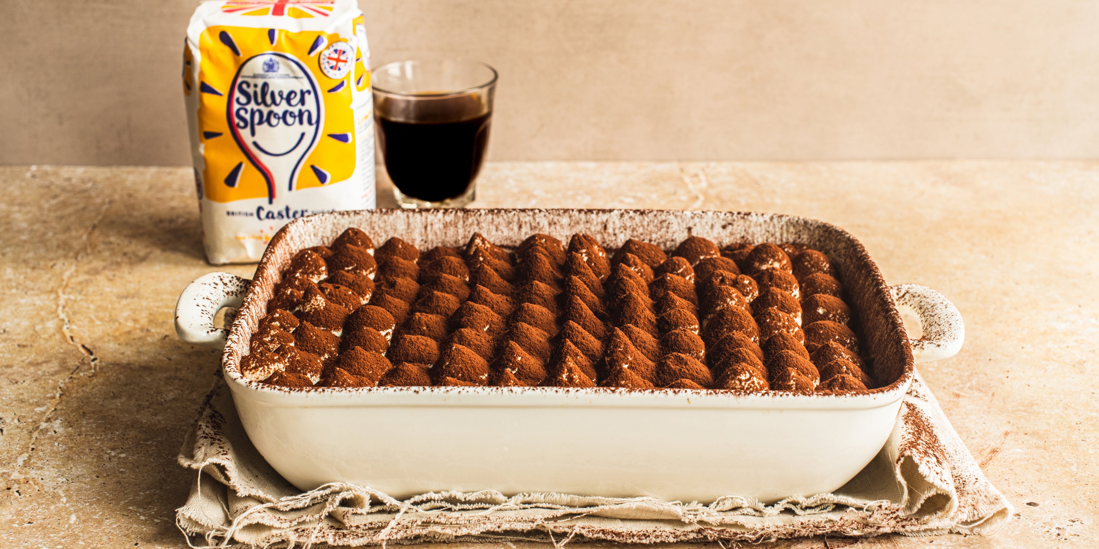 Close up of a dish full of Vegan Tiramisu dusted with cocoa powder, with a bag of Silver Spoon caster sugar and a glass of coffee in the background