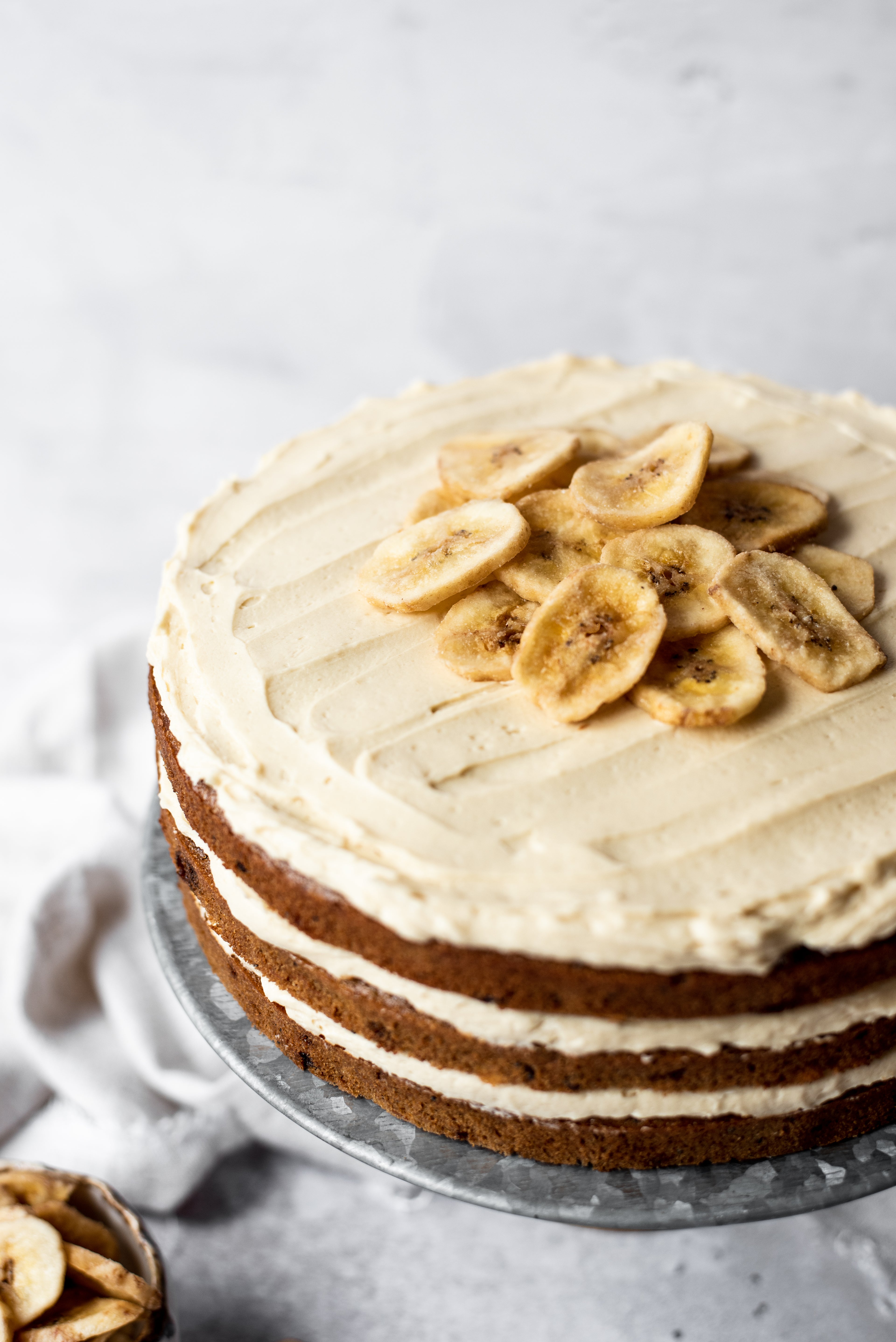 Close up of banana layer cake with 3 layers on cake stand topped with buttercream and banana chips