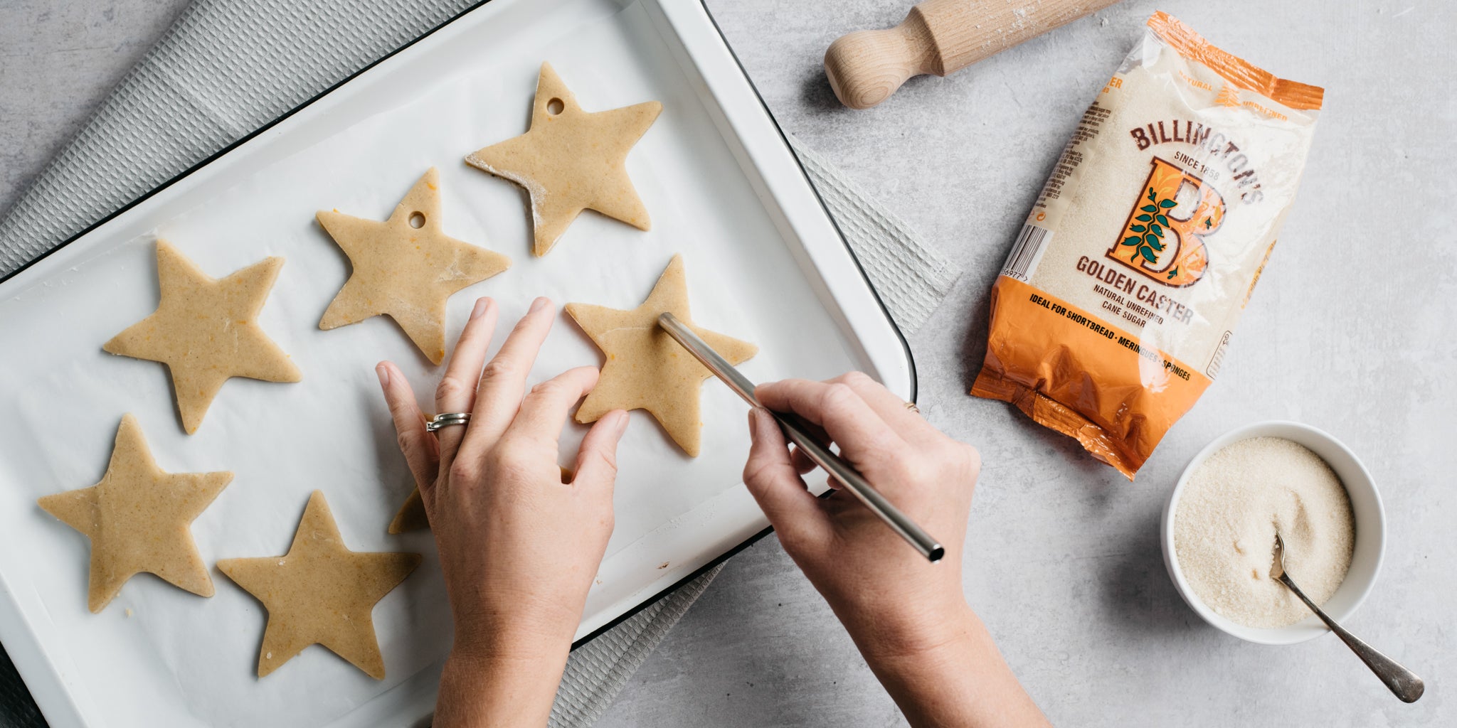 Orange & Ginger shortbread stars being hand decorated, punching a hole into the dough to hang with ribbon once baked. Next to a bag of Billington's golden caster sugar