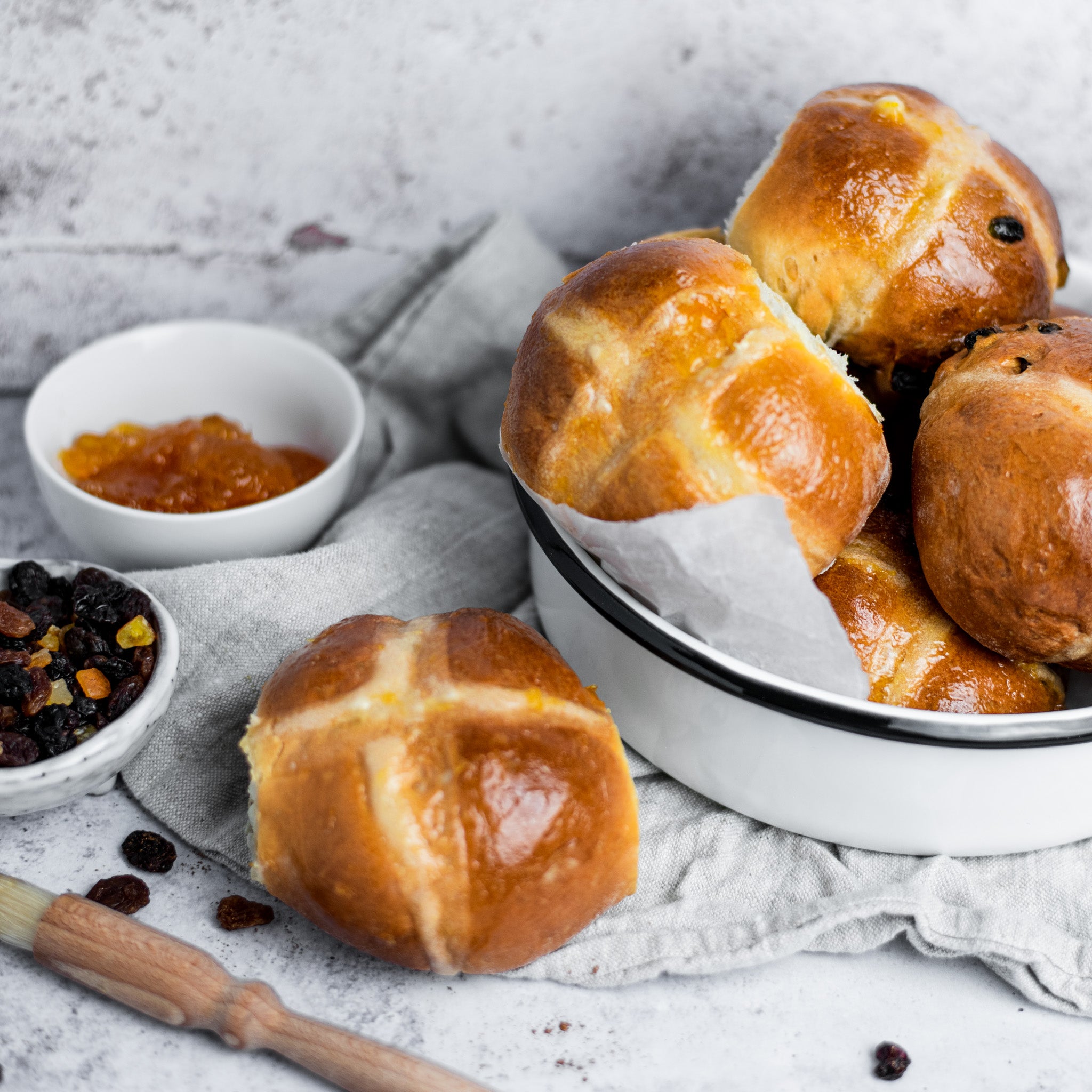 Bowl filled with hot cross buns next to a bowl of sultanas and apricot jam