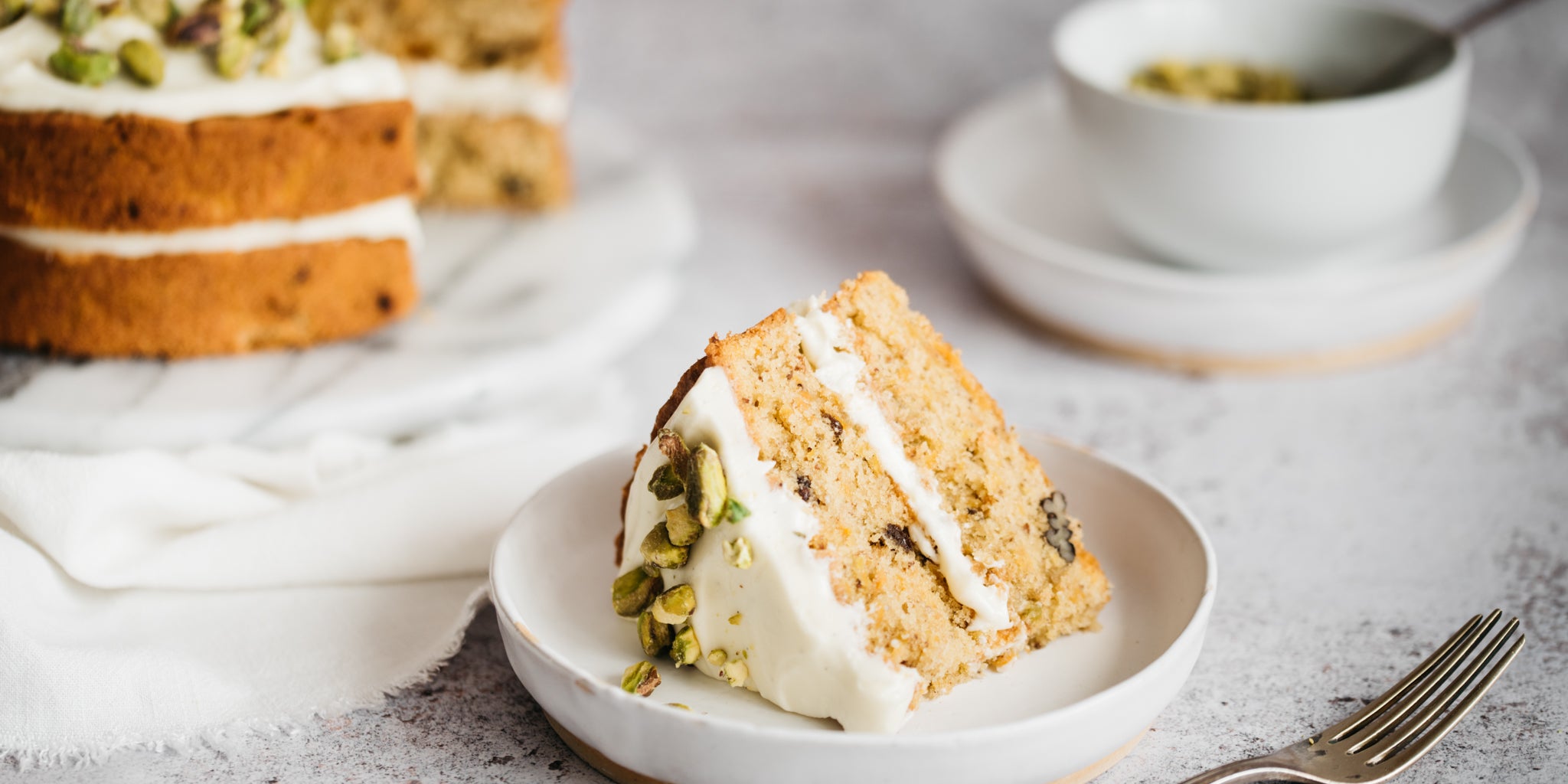 Slice of cake on a small white plate with full cake in the background. Fork in the foreground