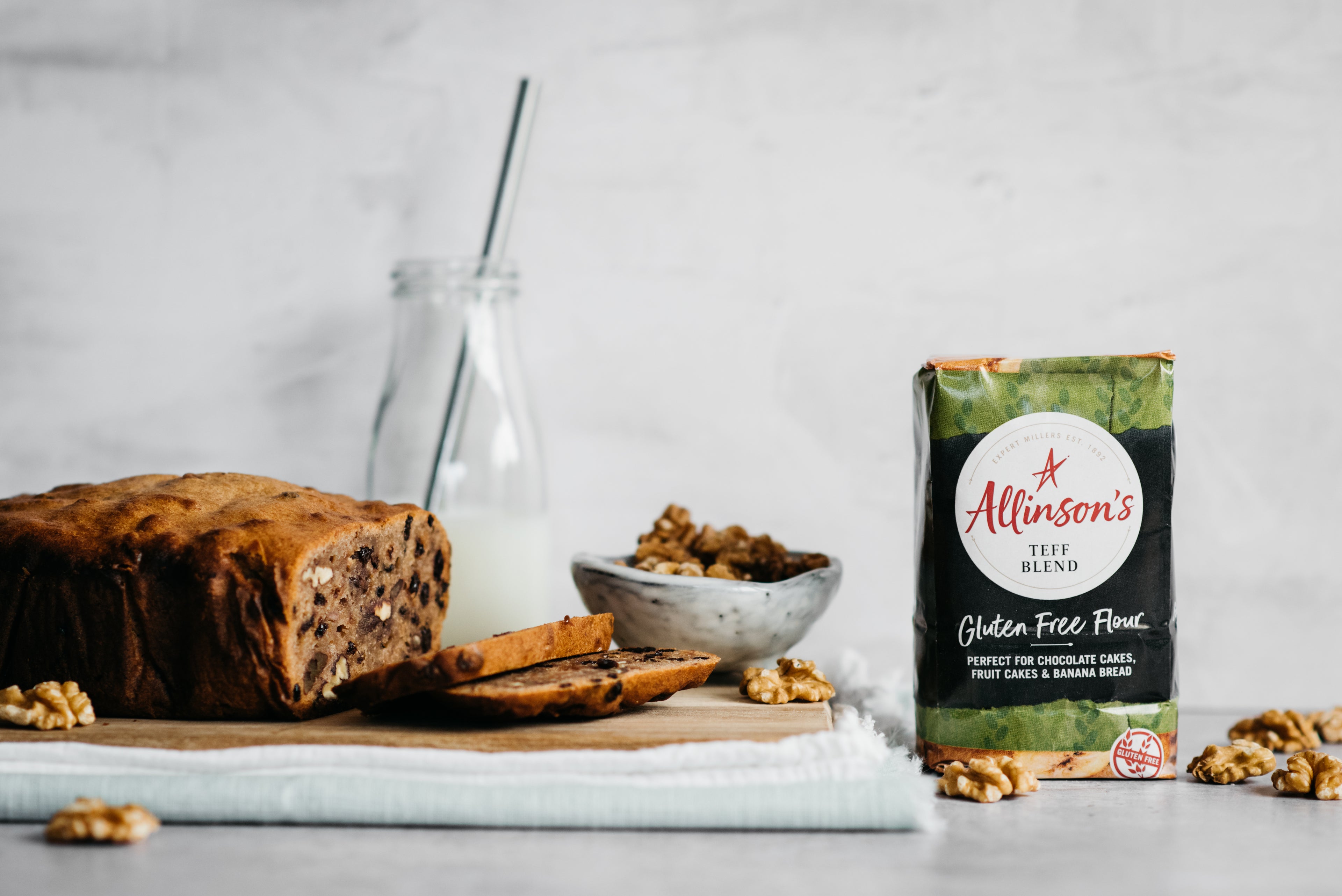 Banana bread, slice cut infront, bowl of walnuts, flour pack and milk bottle in background