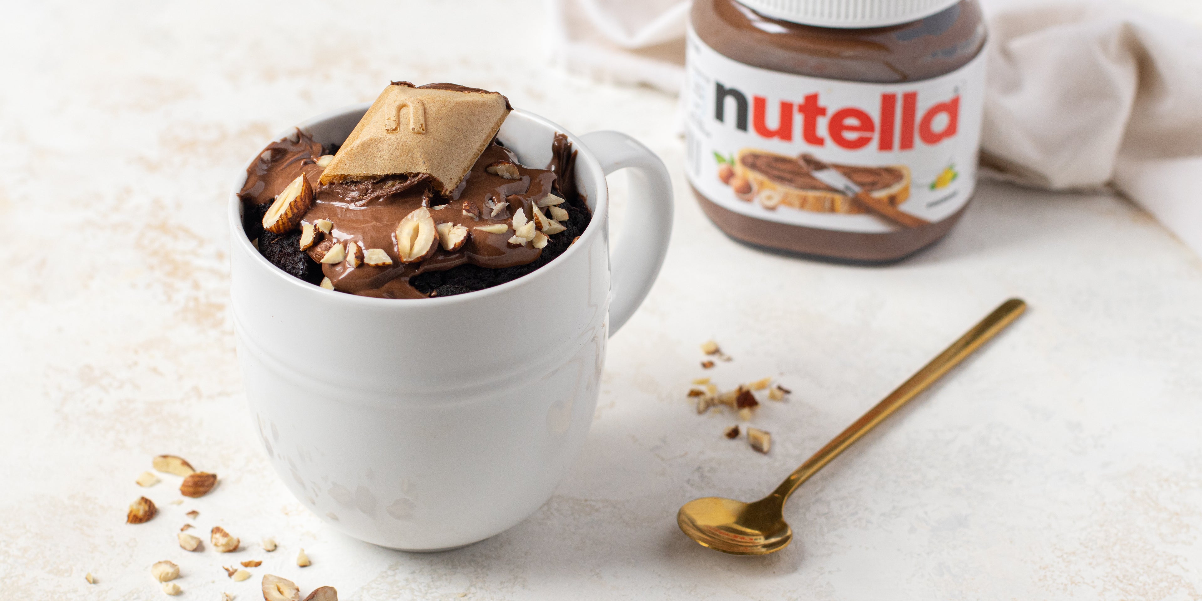 Chocolate Mug Cake customised with Nutella, with Nutella spread on top and sprinkled with hazelnuts. In the background there is a jar of Nutella next to a spoon