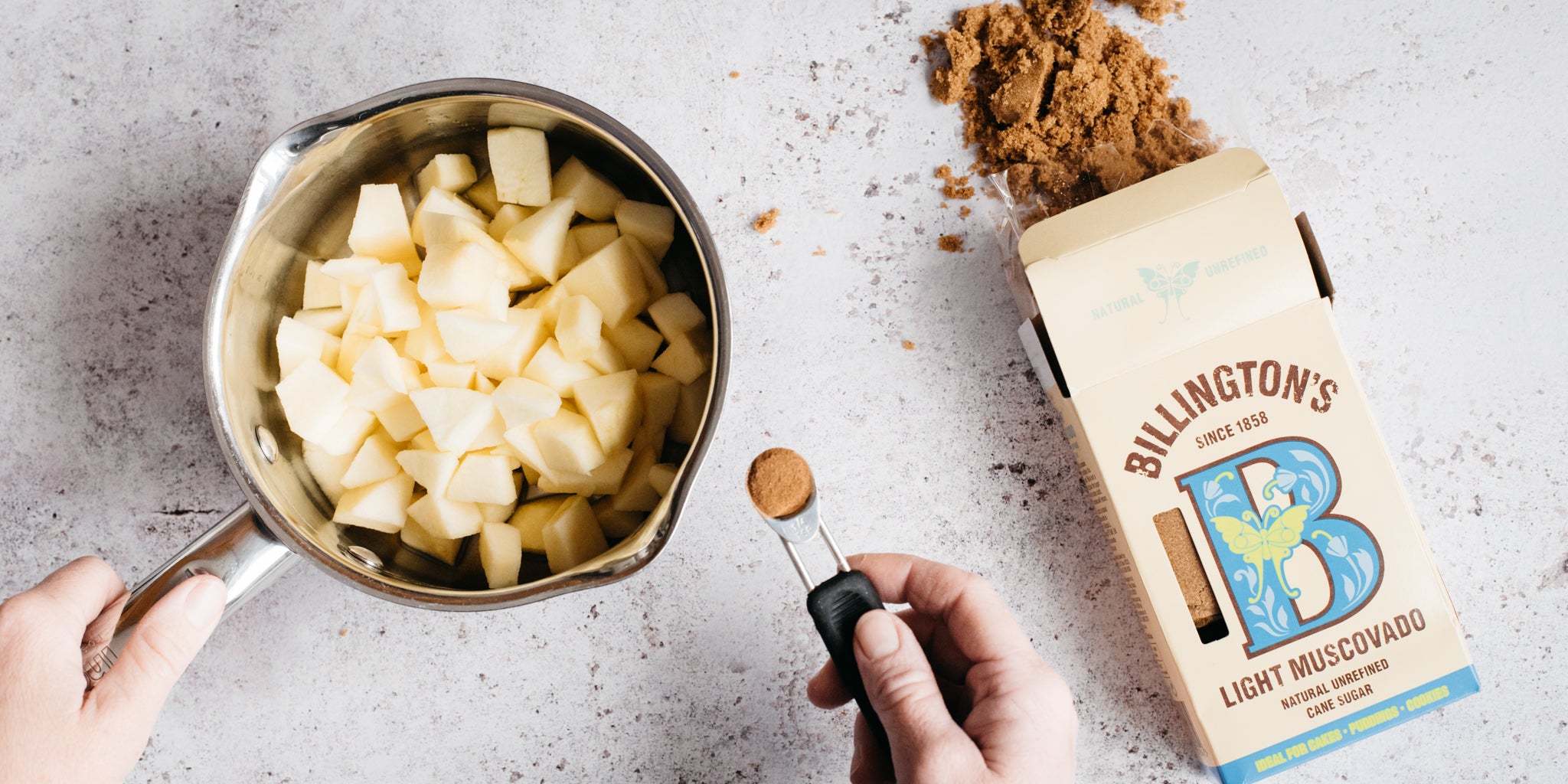 Apples in a saucepan with a teaspoon of cinnamon being added. Open pack of sugar beside it