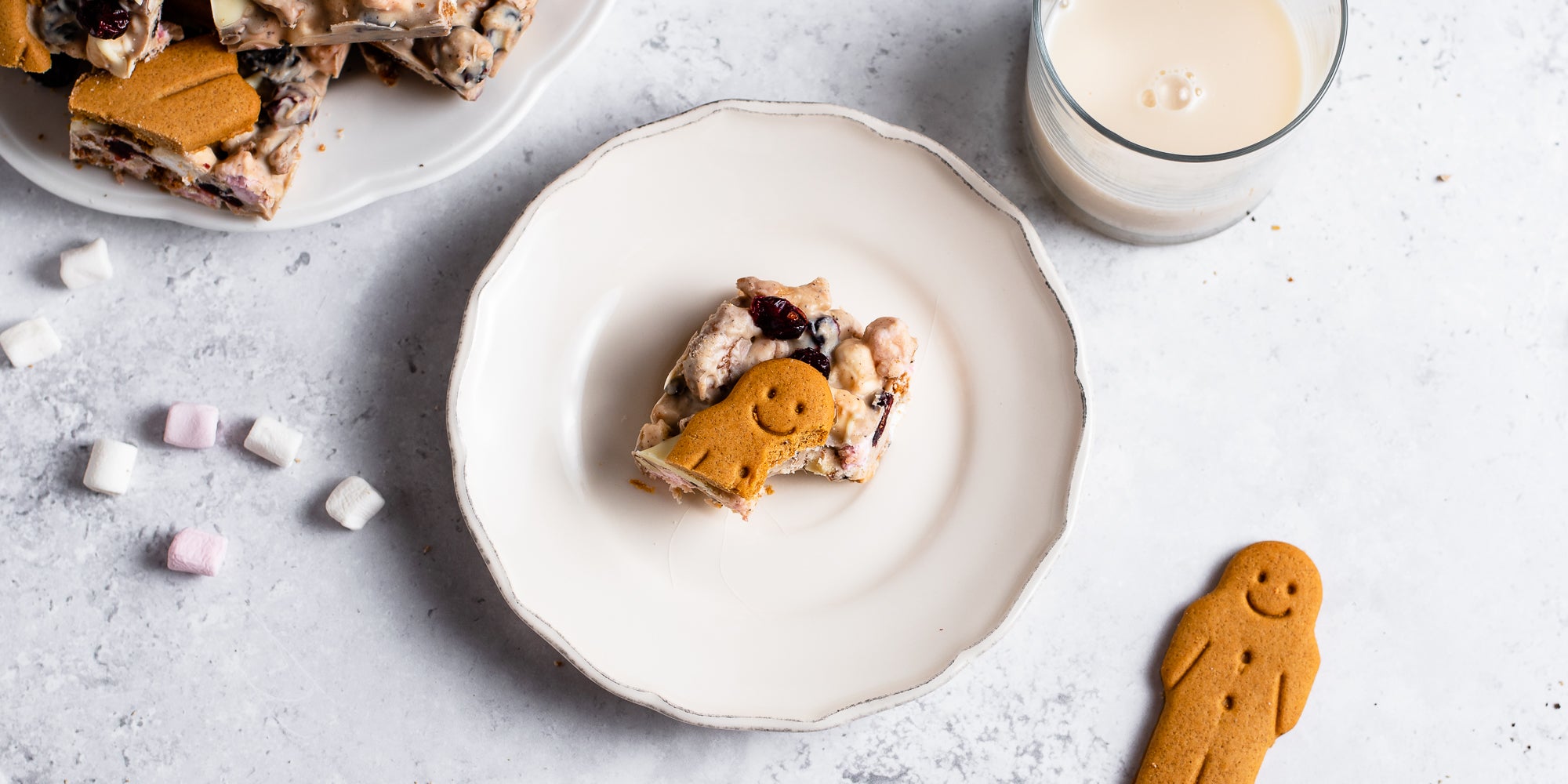 Half eaten slice of White Chocolate & Cranberry Rocky Road on a plate, next to a gingerbread man and mini marshmallows