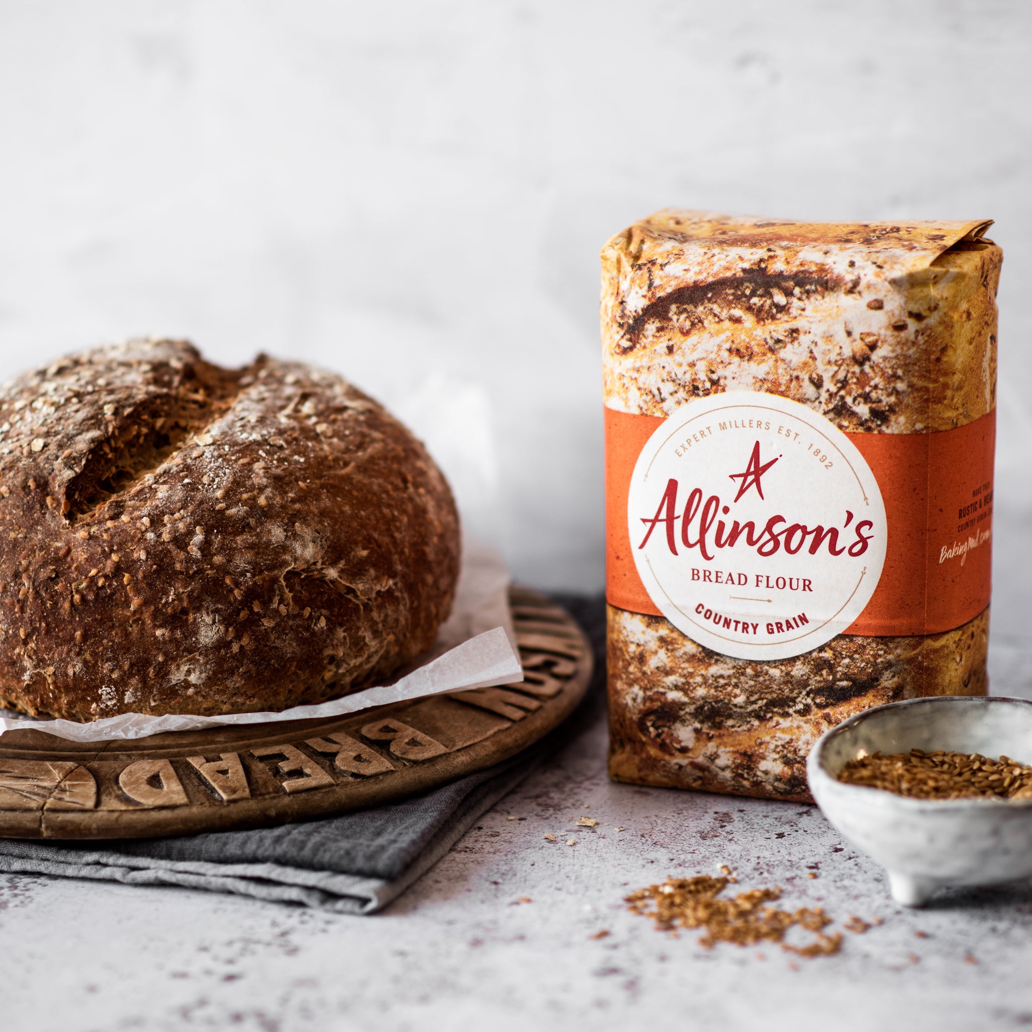 Round loaf of bread on bread board with bowl of seeds and bread flour