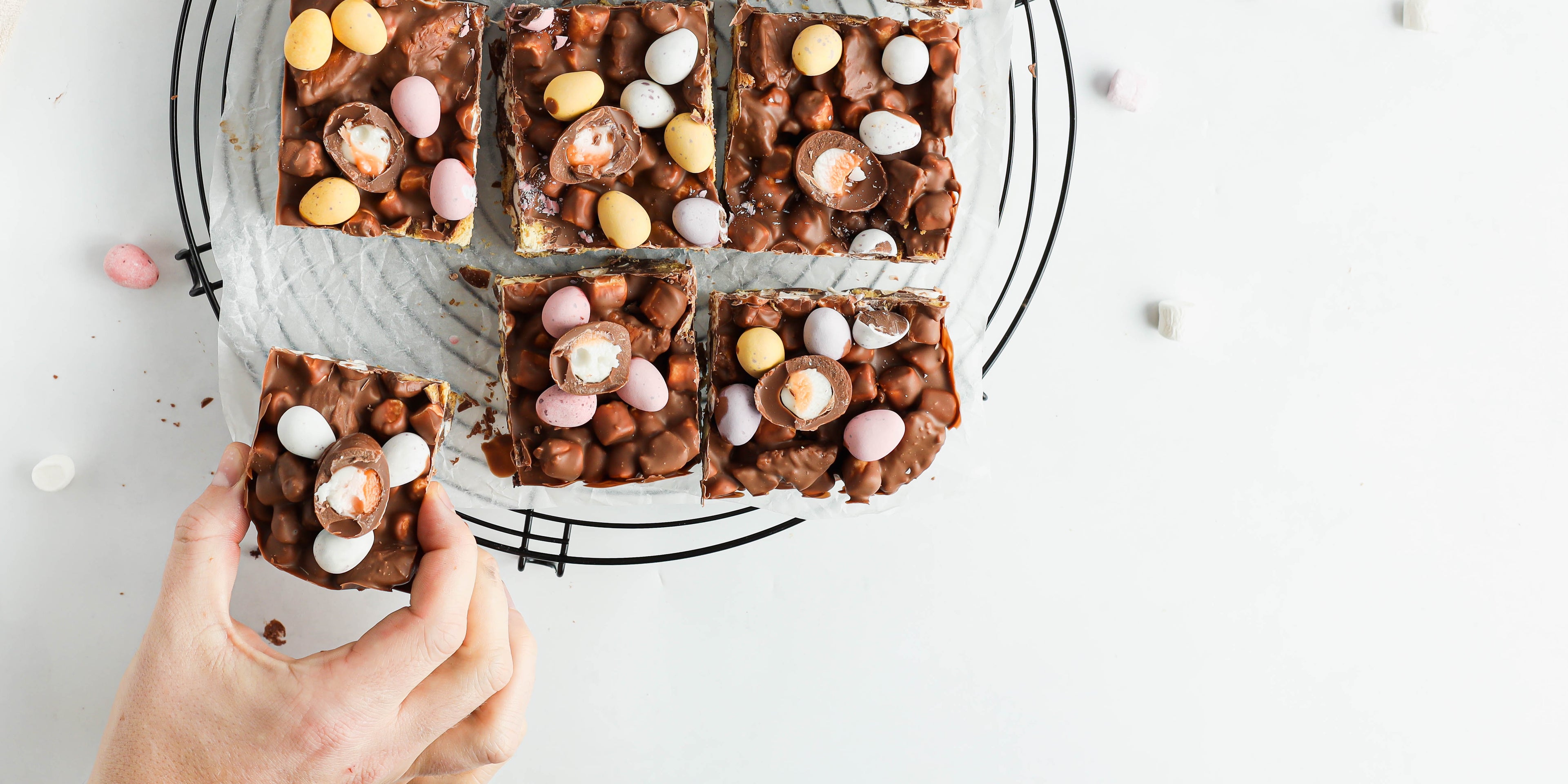 Easter Egg Rocky Road with a hand reaching for a slice, on a wire cooling rack. Topped with creme eggs, mini eggs and marshmallows