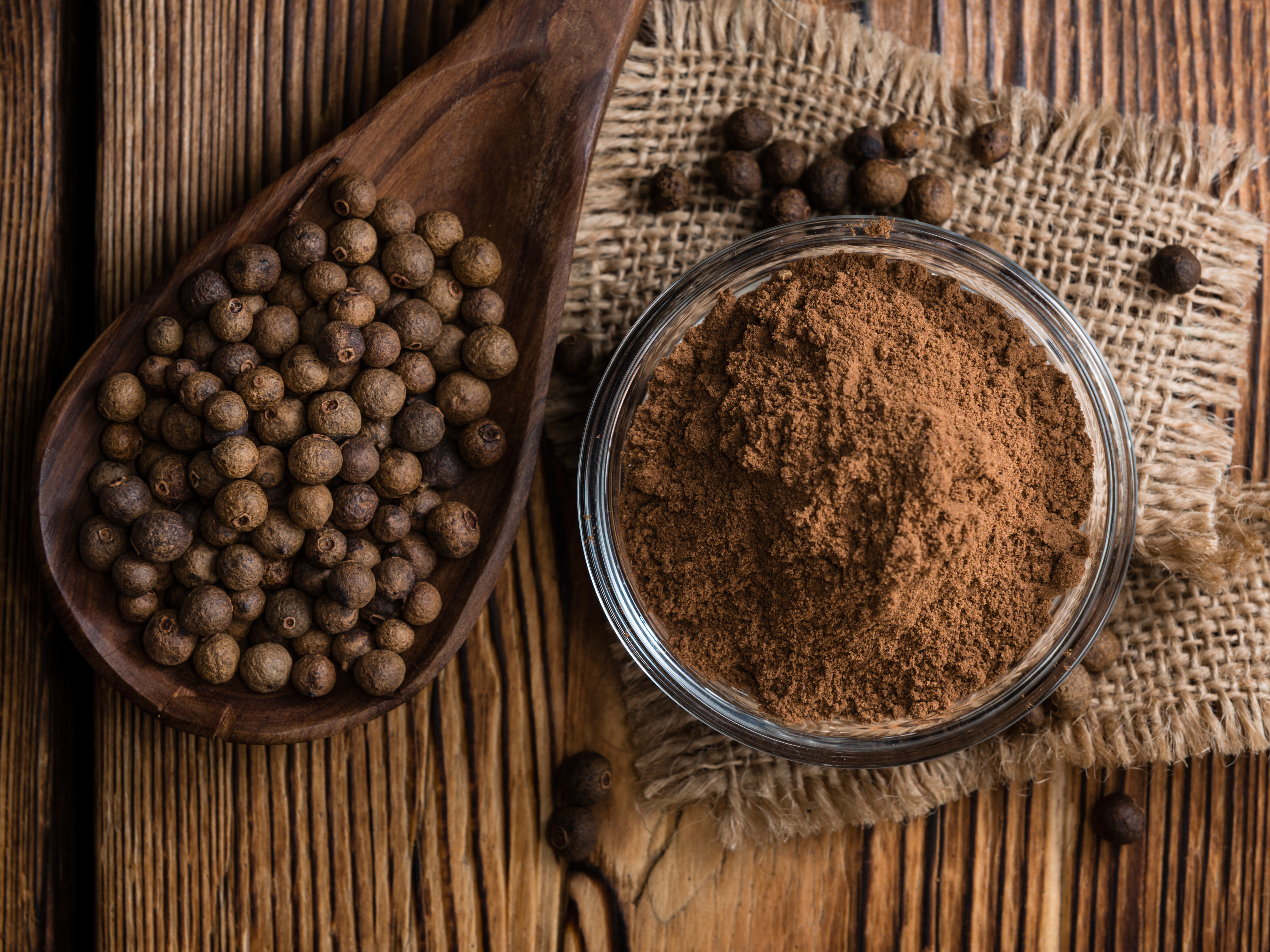 Wooden spoon with whole allspice berries and bowl full of ground allspice