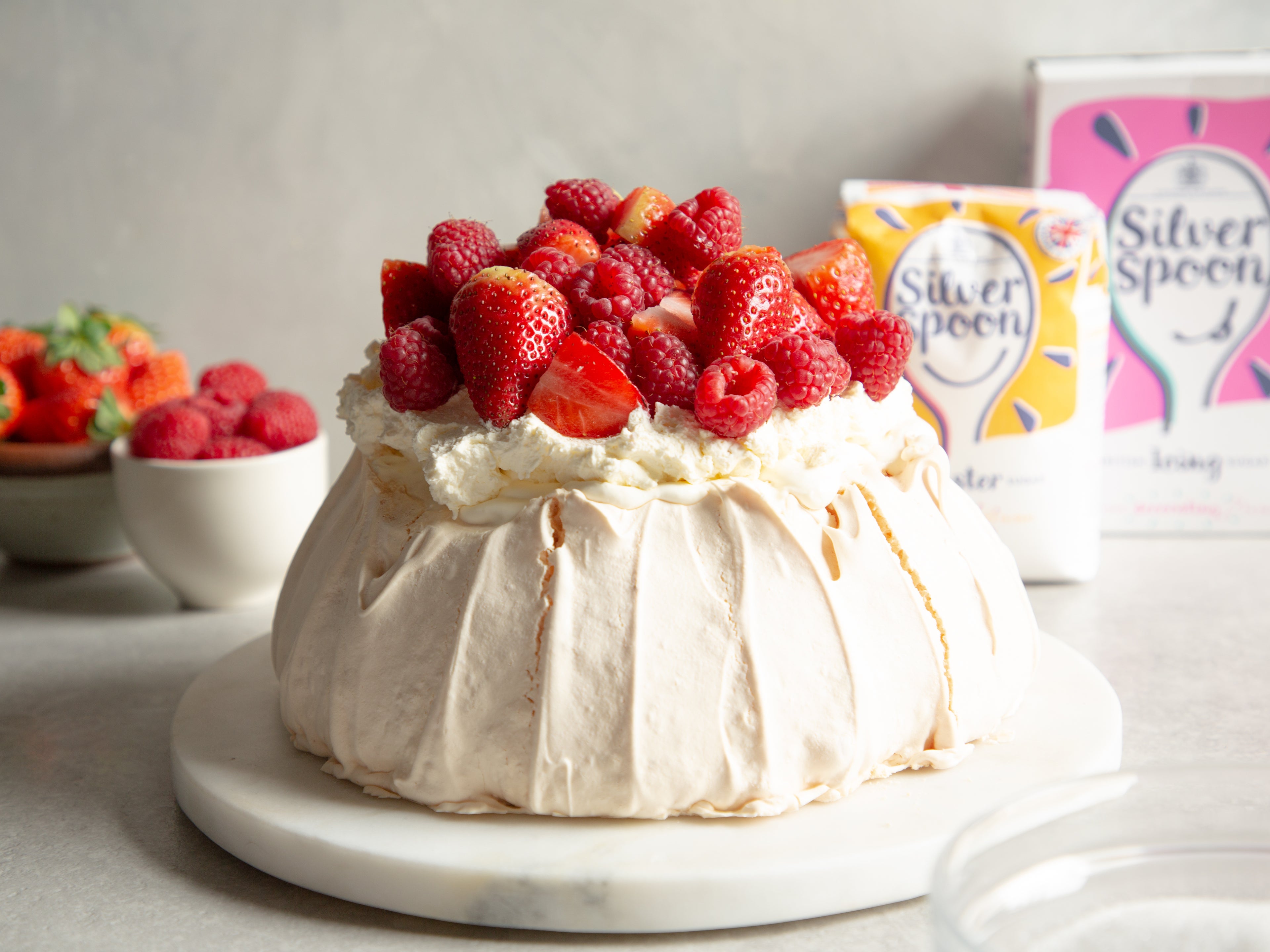Close up of Strawberry Pavlova topped with fresh summer berries, with a box of Silver Spoon Caster and Icing sugar in the background