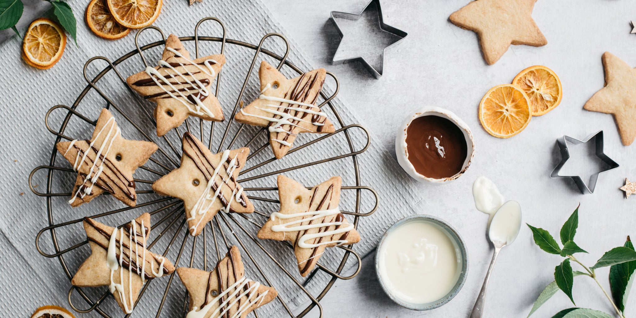Top view of Orange & Ginger shortbread stars on a wire rack, next to a bowl of melted chocolate, sliced orange and cookie cutters.