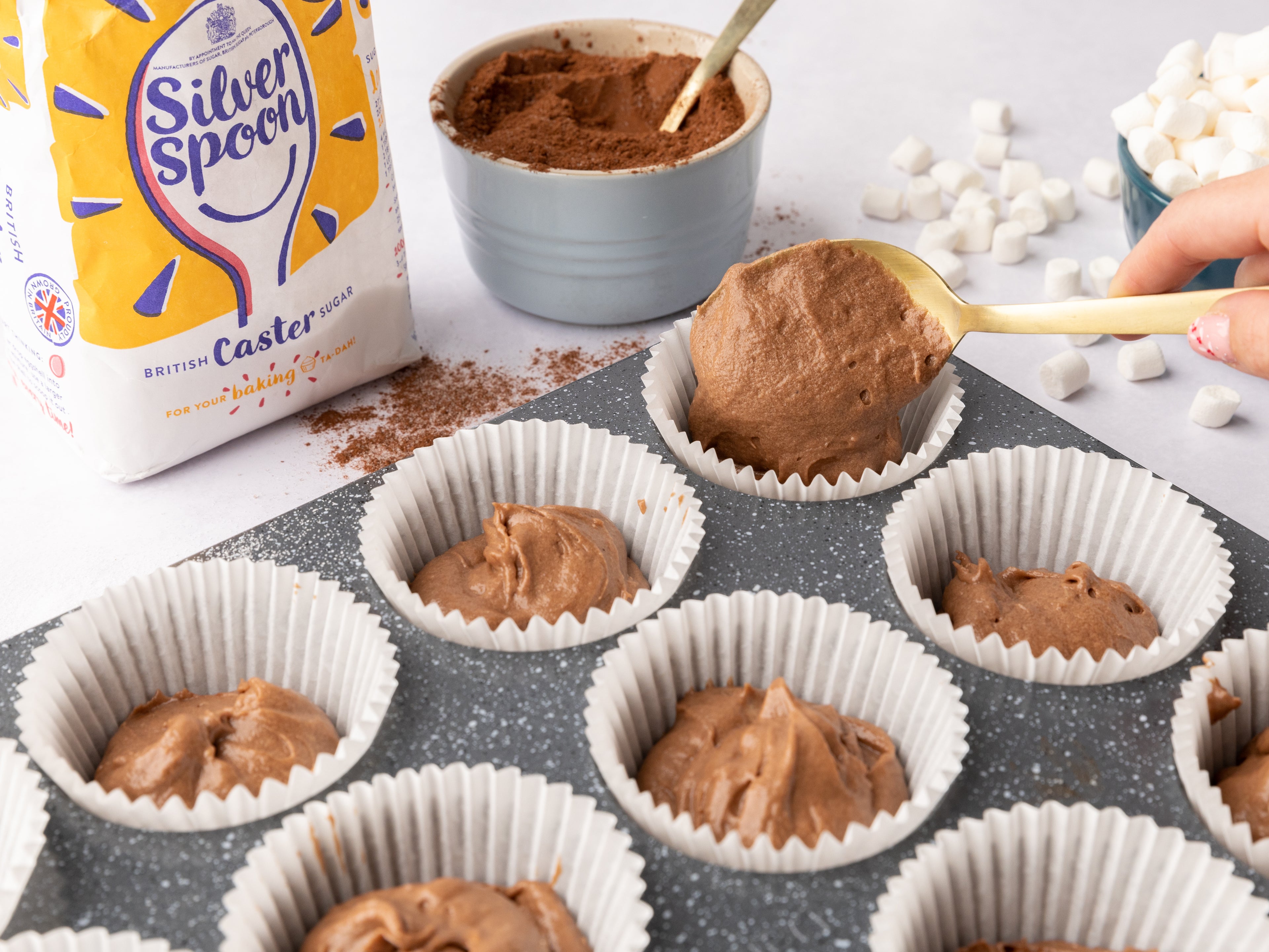 Hand spooning chocolate cake mixture into cupcake cases beside a pack of sugar