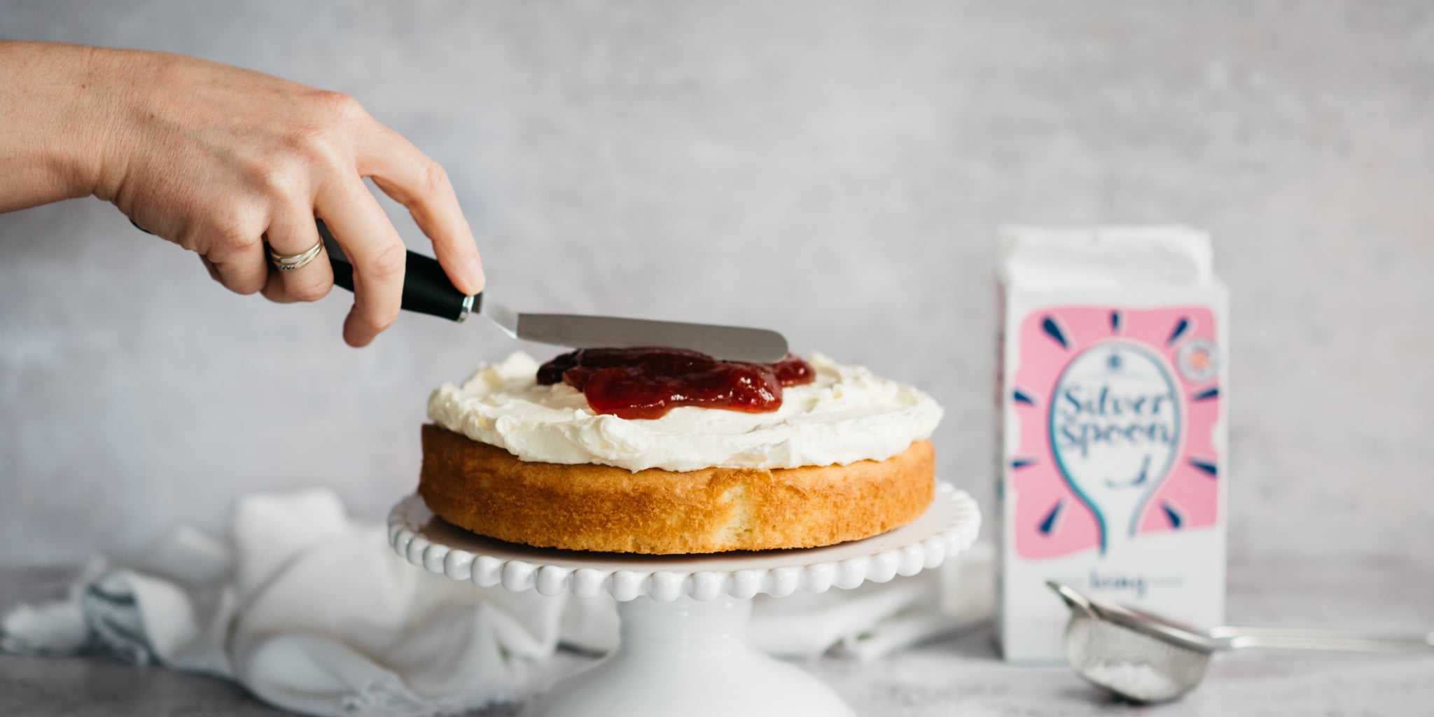 Hand using an offset spatula to spread a layer of jam and cream between the layers of the Gluten Free Victoria Sponge, with a box of Silver Spoon Icing Sugar in the background