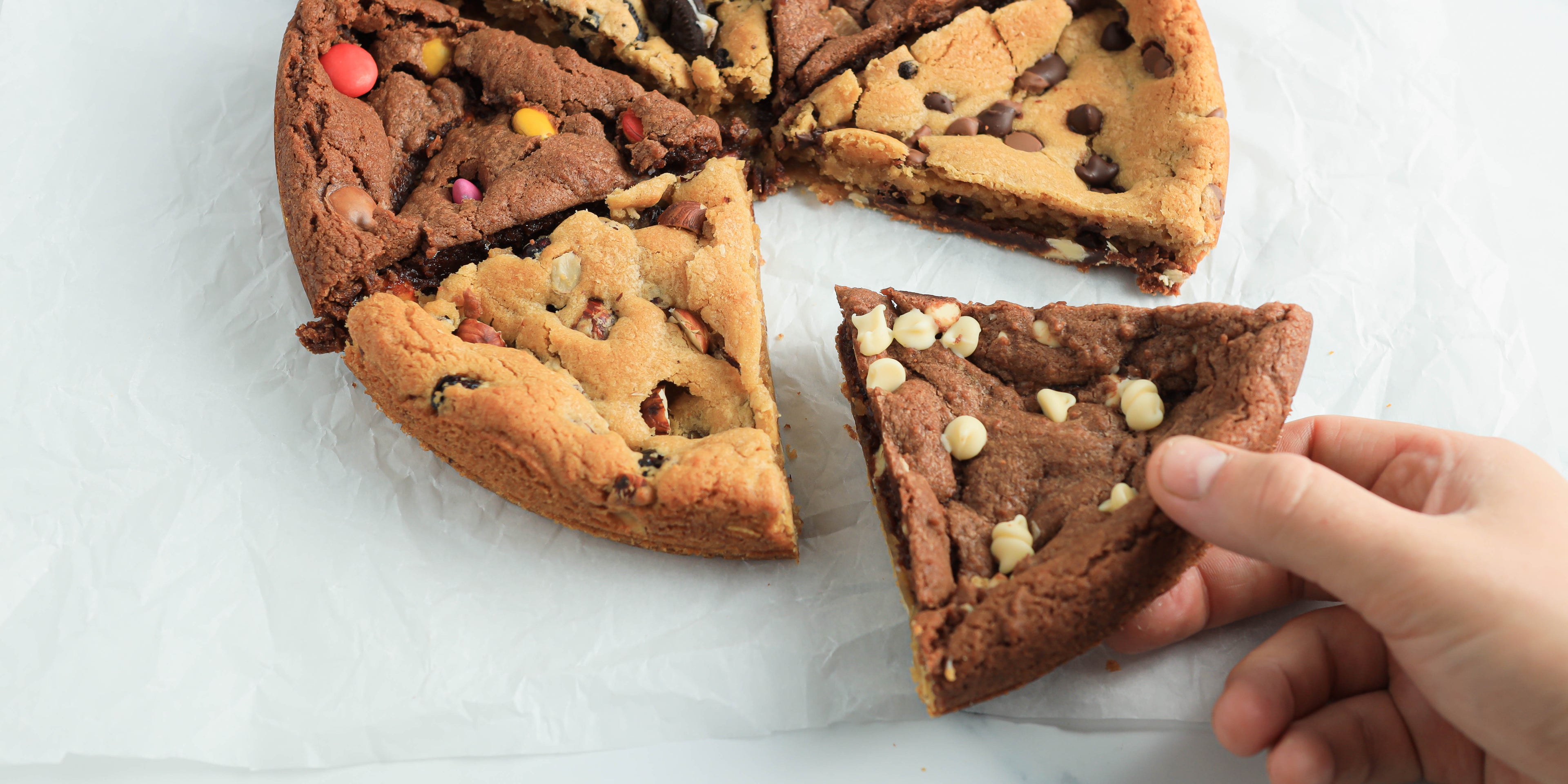 Hand reaching in taking a slice of chocolate cookie with white chocolate chips from a sliced up cookie cake